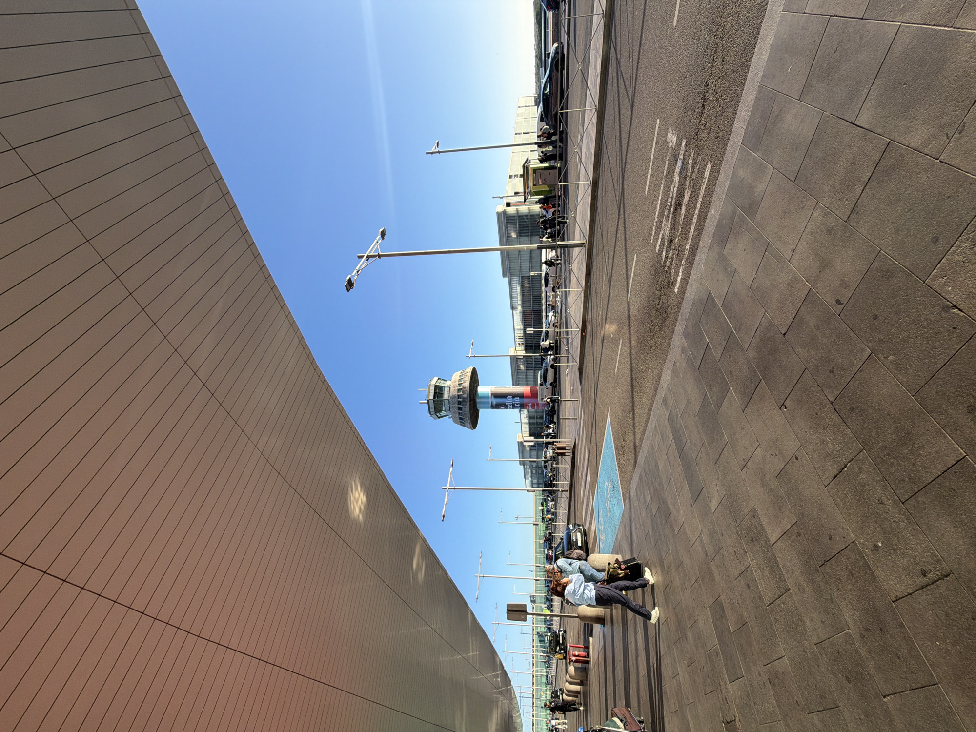The image captures a scene at the El Prat de Llobregat airport in Spain. The architectural elements include a building with a modern, tiled exterior, and the airport's control tower. A street with parked vehicles and lampposts runs parallel to a pedestrian sidewalk. A person is visible on the sidewalk, with luggage nearby. The sky is clear and blue.