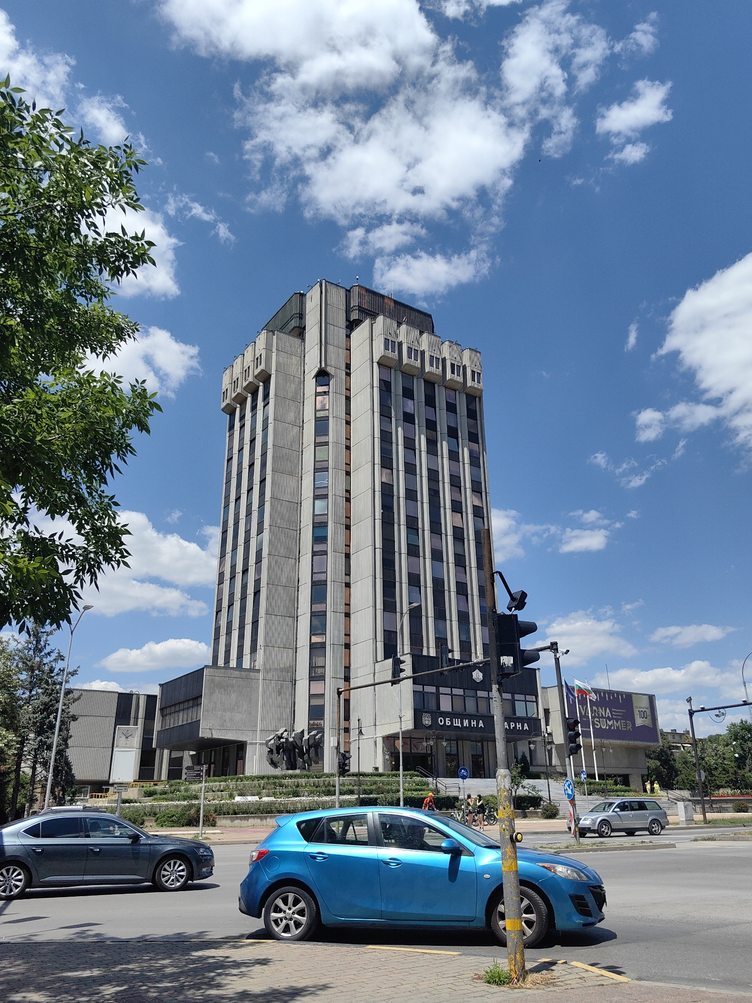 The image captures a street-level view in the city of Varna, Bulgaria, dominated by a tall, Brutalist-style building. The building, likely a civic center or administrative building, features a concrete facade with rows of windows. In front of it, a lower, attached structure has signage that reads "ОБЩИНА ВАРНА" (Municipality of Varna). Green trees line the left side of the frame, with their leaves partially obscuring the sky and the upper portion of the main building.
The street in the foreground is paved with asphalt and marked with white lines. A blue hatchback car is positioned in the right lane, facing towards the viewer, its front right tire visible. To the left of the blue car, a darker colored sedan is driving away from the viewer in the left lane. The sky above is a bright blue, dotted with scattered white cumulus clouds. Traffic lights and street signs are visible on a pole in the middle ground, near the entrance of the building. A Bulgarian flag is flying on a flagpole to the right of the building entrance. In the background, more trees and some smaller buildings are visible, along with a billboard that says "VARNA SUMMER". A few pedestrians and cyclists are seen in the middle ground near the building entrance. The scene appears to be set during daylight hours on a clear day.