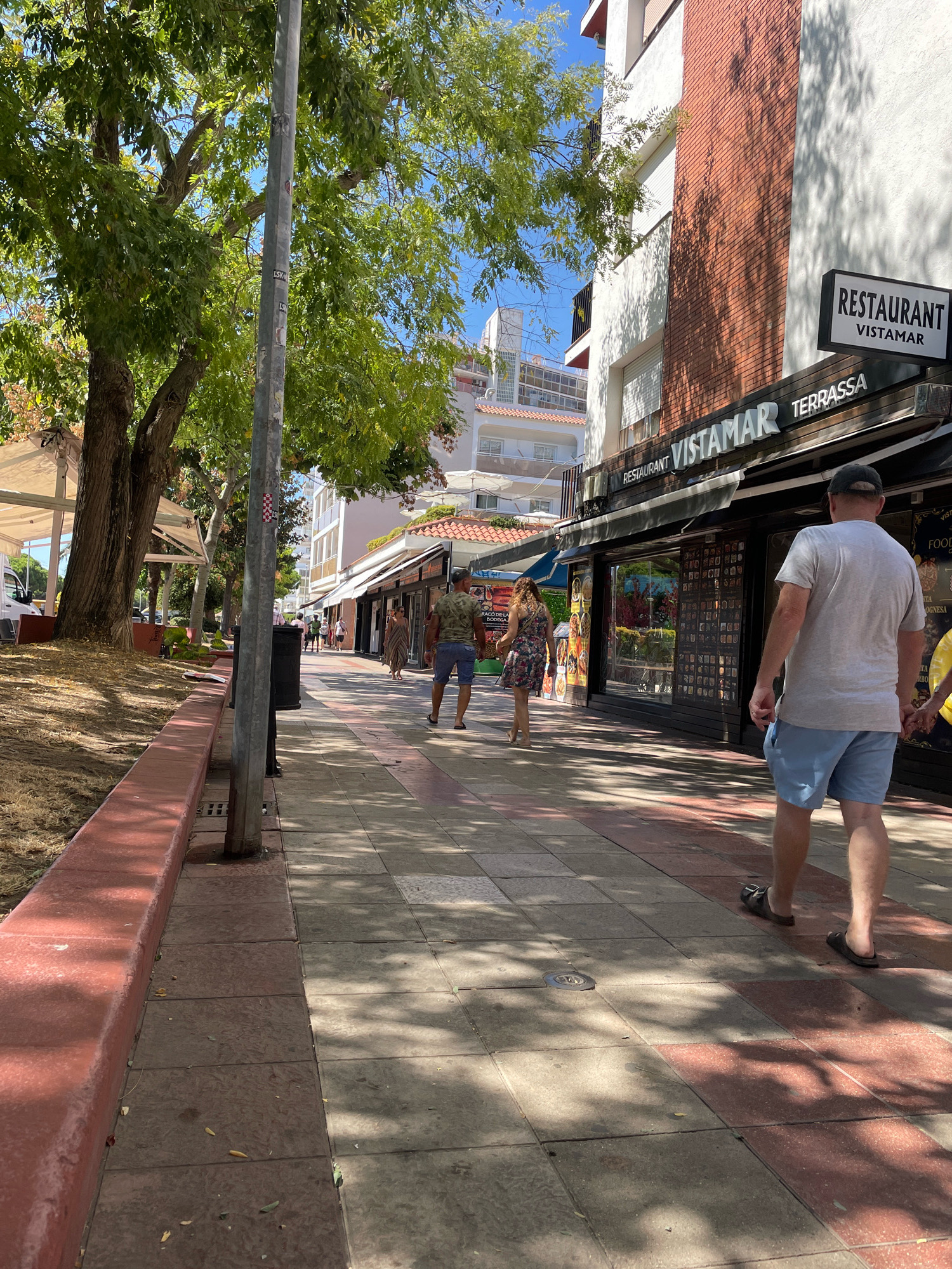 The image depicts a sunlit pedestrian sidewalk in Malgrat de Mar, Spain, likely along a commercial street. A line of trees with green foliage shades the left side of the sidewalk, casting dappled shadows on the ground. The sidewalk itself is paved with square tiles in varying shades of red and grey. A red-painted curb runs along the left edge of the sidewalk.
Several people are visible walking along the sidewalk. In the foreground, a man wearing a grey t-shirt and light blue shorts walks away from the camera, his back to the viewer. He is wearing dark sandals. Further down the sidewalk, a woman in a floral print dress walks away from the camera, and behind her, a man in a patterned t-shirt and blue shorts also walks away. Other figures are further down the sidewalk, appearing as smaller shapes due to distance.
On the right side of the sidewalk are the storefronts of businesses. One building features a facade with red brick accents and white stucco. A prominent sign reads "RESTAURANT VISTAMAR TERRASSA". The windows of the establishments display menus and products. The sky is visible as a clear blue. The overall impression is of a pleasant, sunny day in a busy town.