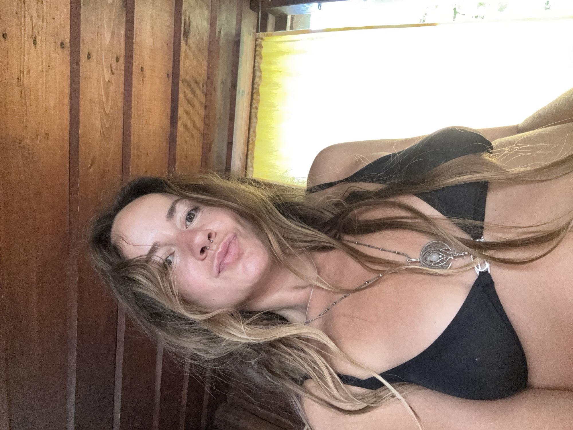 A woman is lying down, facing the camera, with her head tilted and her hair spread out. She is wearing a black bikini top and a silver necklace with a pendant. The background consists of wooden walls and a yellow curtain. The lighting suggests it might be daytime, with sunlight filtering in from behind the curtain. The woman's expression is playful, with her lips pursed. There is no visible text in the image. The overall scene appears to be casual and relaxed, possibly in a rustic or beach-like setting.