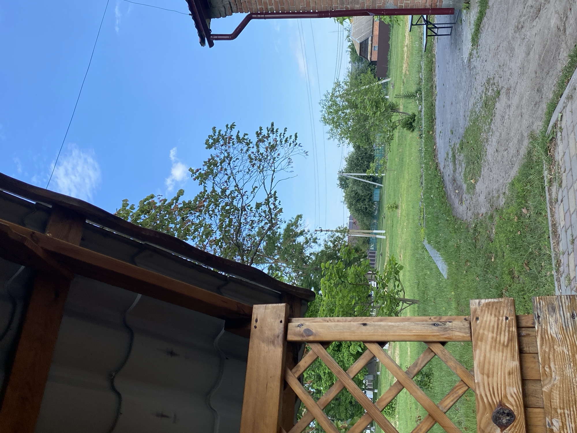 The image depicts an outdoor scene with a wooden gazebo in the foreground. The gazebo has a sloped roof covered with grey corrugated metal and wooden beams. To the right of the gazebo, a brick building with a maroon gutter is visible. Beyond the gazebo, a paved area leads to a grassy yard. Several trees are scattered across the yard, with a few houses and power lines visible in the background under a clear blue sky with sparse clouds. A picnic table with dark metal legs is positioned near the brick building. The ground is a mix of dirt, grass, and paving stones.