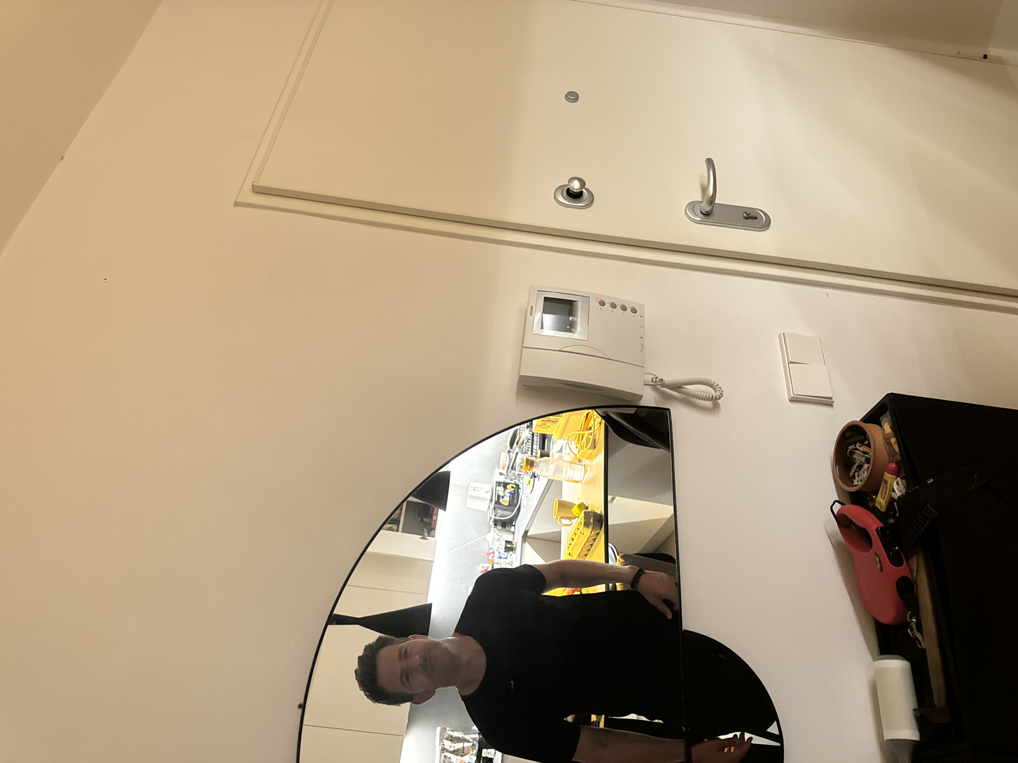 In a residential hallway, a man in a black t-shirt stands before a large, arched mirror, his reflection filling a portion of the frame. The mirror reveals a glimpse of a kitchen with countertops and cabinets, suggesting the photo is taken near the entrance to a home. To the right of the mirror, a cream-colored intercom system is mounted on the wall. Adjacent to the intercom and slightly below, a white light switch is visible. Further to the right, a closed, cream-colored door with silver hardware, including a doorknob and a keyhole, occupies the majority of the right side of the image. In the foreground, on a black surface, a red retractable dog leash, a roll of toilet paper, a bowl filled with keys, and a yellow tube are arranged. The overall scene is well-lit, indicating an indoor setting with artificial lighting. The architectural style and the presence of an intercom system are consistent with modern apartment buildings, and the detail about Murcia, Spain, is not visually ascertainable from the provided image.