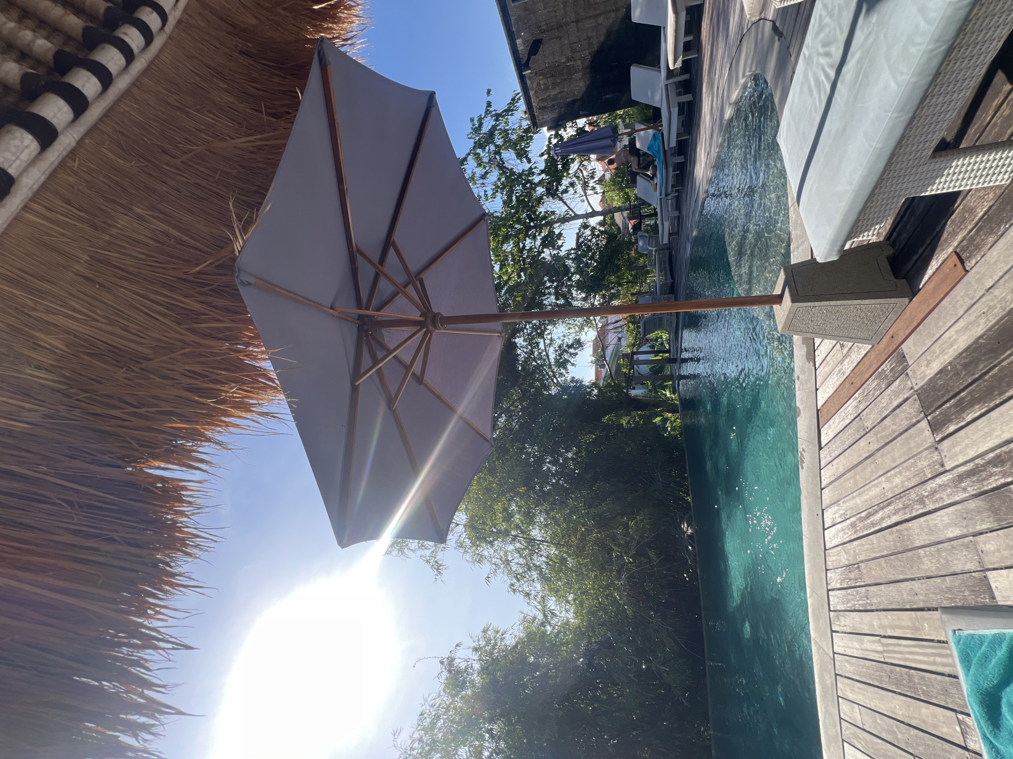 The image shows an outdoor scene, likely a vacation or resort setting. A grey umbrella is centrally located, partially blocking the view of the sun and sky. A thatched roof structure is visible in the top-left corner. There is a pool adjacent to a wooden deck with lounge chairs. In the background, trees and foliage obscure the view, with a few buildings visible. Based on the image's origin and the location tag, the scene is likely in North Kuta, Indonesia. The scene appears to be unoccupied at the moment of capture.