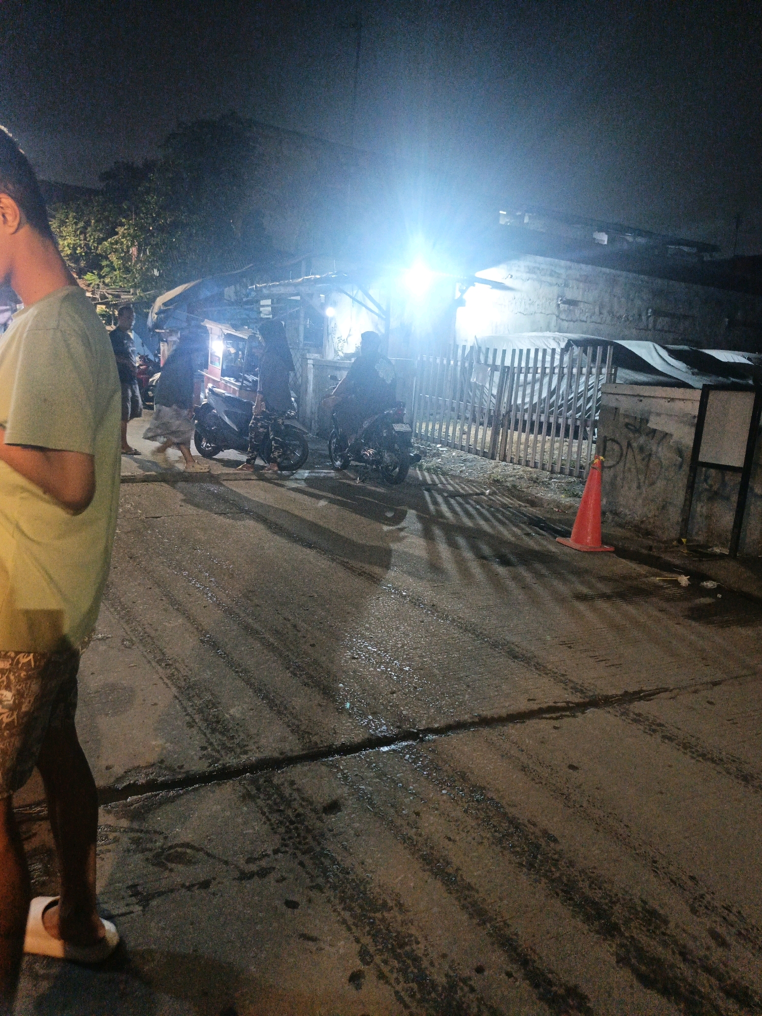 The image depicts a nighttime scene on a street in North Jakarta, Indonesia. The foreground features the partial view of a person wearing a light green t-shirt and patterned shorts, with one foot in a white sandal. The street surface is asphalt, wet and marked with tire tracks. In the midground, several motorcycles are visible. One person is on a motorcycle that appears to be stopped, while another person stands beside it. Further back, under a dimly lit structure that might be a market stall or informal shop, more people are gathered. A bright, overexposed light source illuminates the area from above and to the left, creating harsh shadows. To the right, a red traffic cone is placed on the ground near a wall with graffiti. The background consists of more structures and trees against a dark night sky. The overall impression is of a busy, possibly informal, urban environment at night.