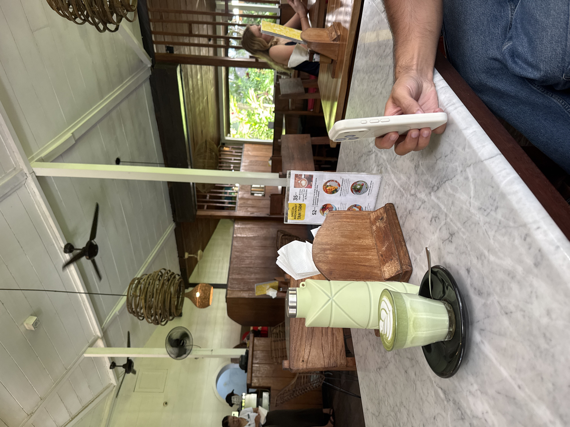 This image, taken indoors in North Kuta, Indonesia, shows a marble-top table in a restaurant. A person's hand holding a white smartphone is visible in the upper right corner. The table holds a glass of green matcha latte on a black saucer with a spoon, a green water bottle, a wooden tissue box with tissues, and a menu. Other people can be seen seated at tables in the background, with a view of greenery outside. The restaurant has a high ceiling with wooden beams, ceiling fans, and woven pendant lights.