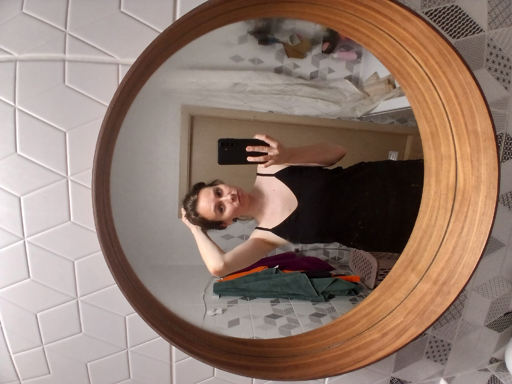 A woman is taking a selfie in a bathroom mirror. She is wearing a black spaghetti strap dress and has her hand in her hair. The bathroom has white geometric tiles on the wall behind her and a patterned tile backsplash around the mirror. There are several towels hanging on a rack to the left of the mirror, as well as a laundry basket. The mirror has a wooden frame.