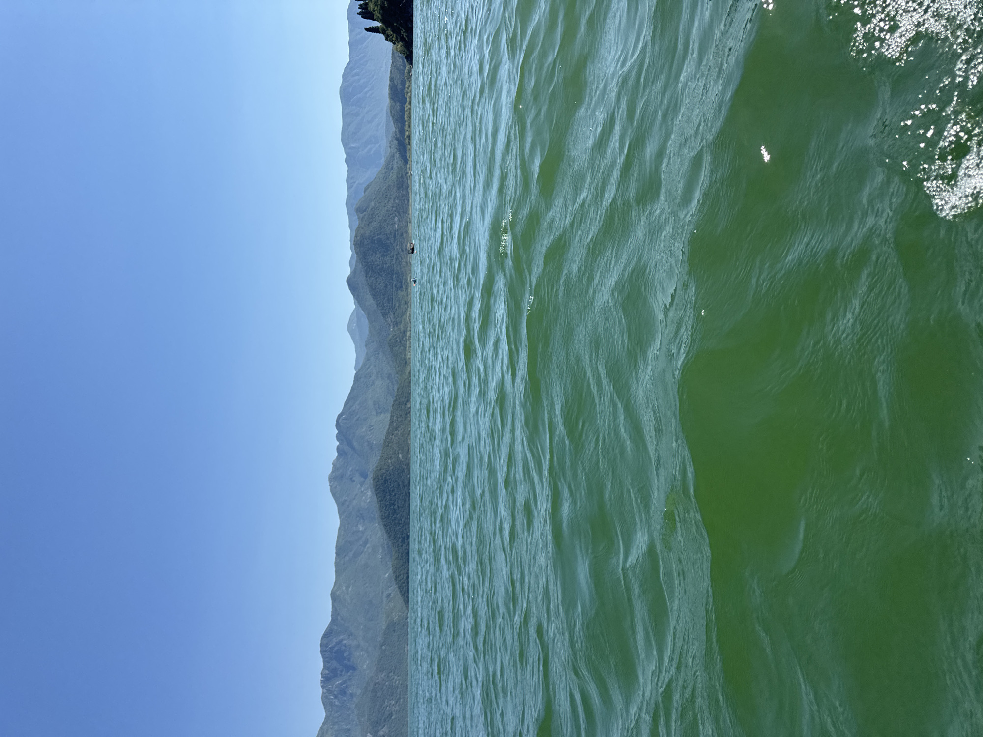 The image depicts the city of Kruševica, Montenegro, from a body of water, presumably a lake or river. The water is choppy and green, with some foam or bubbles visible on the right side. On the left side of the image, mountains rise from the water's edge, covered in green trees. The sky is blue and clear.
