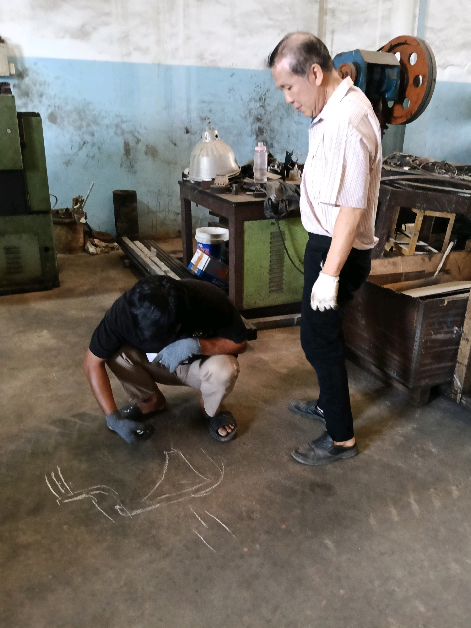 In the city of Belimbing, Indonesia, two men are observed in what appears to be a workshop or industrial setting. One man, wearing a black t-shirt and khaki pants, is crouching on the concrete floor, drawing with white chalk. The drawing on the floor depicts a schematic or diagram. He is wearing grey gloves on his hands. The second man, an older individual with short dark hair, stands nearby, wearing a light-colored, pinstriped collared shirt, black pants, and black shoes. He is also wearing white gloves on his hands and looking down towards the chalk drawing. The environment shows industrial machinery, workbenches, and various tools and materials scattered around, suggesting a functional workspace. The walls are painted blue and white, with visible signs of wear and dirt.