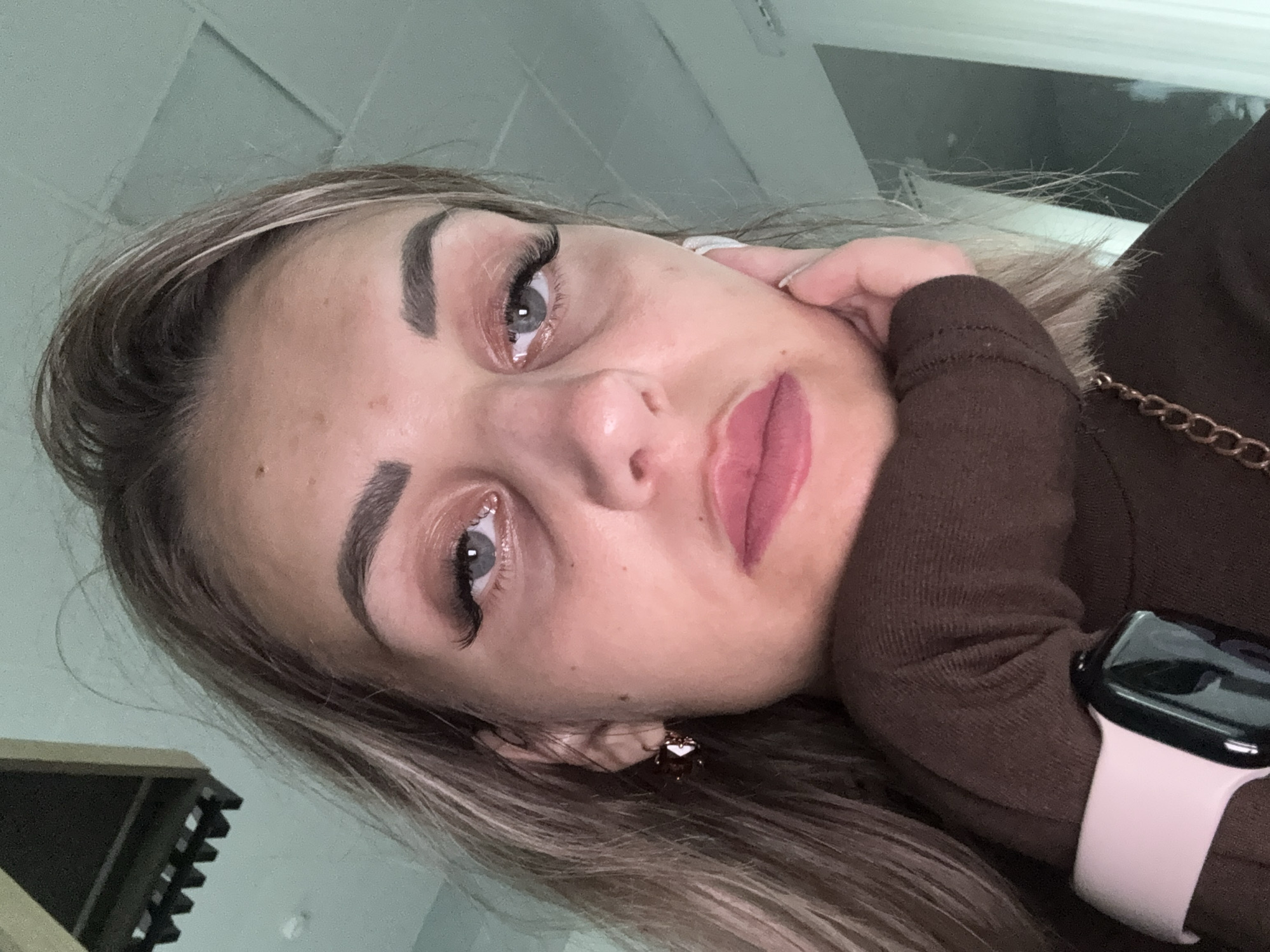 A close-up, slightly overhead, indoor selfie captures a young Caucasian woman in a brown turtleneck. She has blonde hair, styled with some strands loose around her face, and is wearing makeup including eyeshadow and false eyelashes. A large earring with a reddish-brown stone is visible in her left ear. Her left hand rests on her cheek, supporting her head. A light pink smartwatch is on her left wrist. The background is out of focus, appearing to be an office or indoor space with a light-colored ceiling and wall. There are no discernible landmarks indicating the city of Yuzhno-Sakhalinsk, Russia.