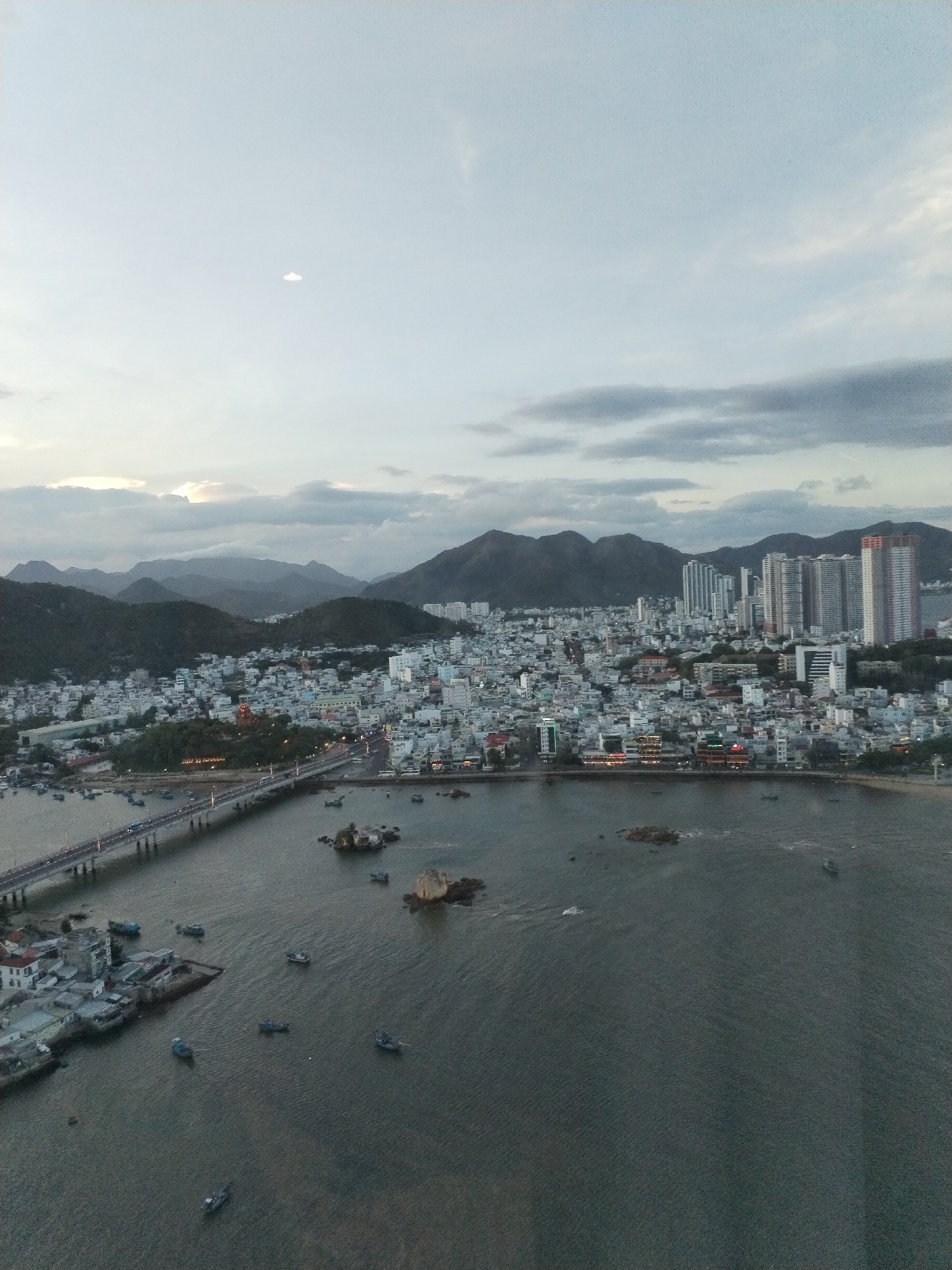 The image depicts a cityscape with a bridge spanning a body of water. In the background, mountains rise beyond the dense urban area, which consists of a mix of low-rise buildings and high-rise towers. The sky is overcast with visible cloud cover. Several small boats are scattered on the water's surface. The location is Bac Nha Trang Ward, Vietnam.