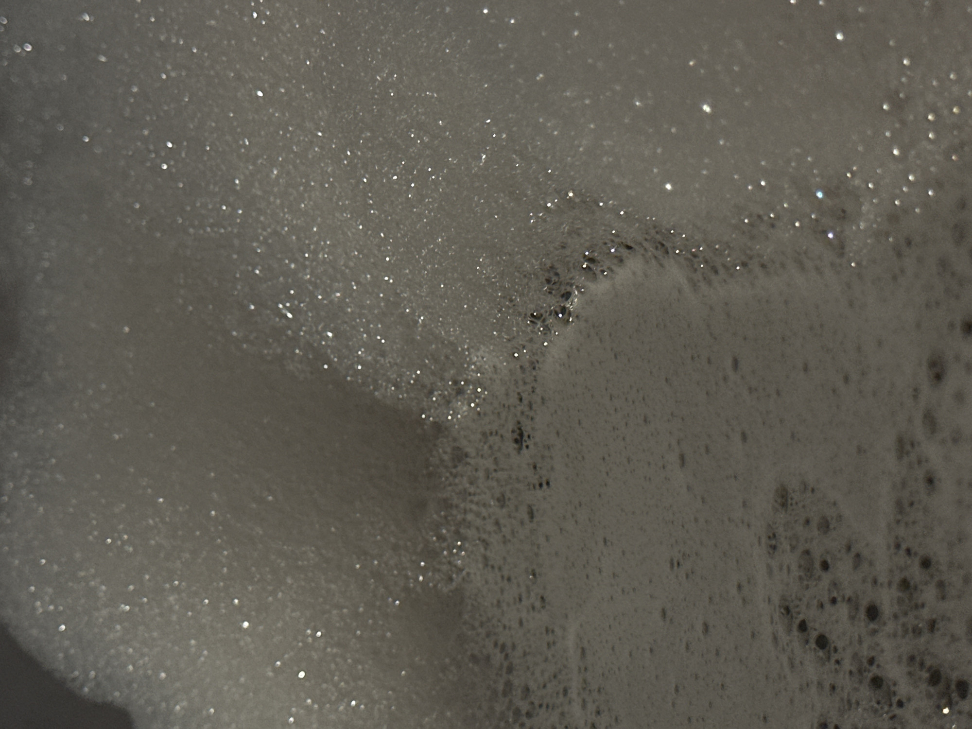 The image is a close-up, abstract photograph of soap suds. The suds are white and foamy, with many small bubbles reflecting light. The lighting suggests the suds are illuminated from above and slightly to the left, creating highlights and shadows. The texture is soft and airy, with a sense of depth created by the varying sizes and densities of the bubbles. The background is out of focus and appears to be a dark, flat surface. There are no people or discernible objects within the frame, only the foamy substance. The image does not contain any information or visual cues that would allow for its localization within Sofia, Bulgaria, or any other specific geographical location.