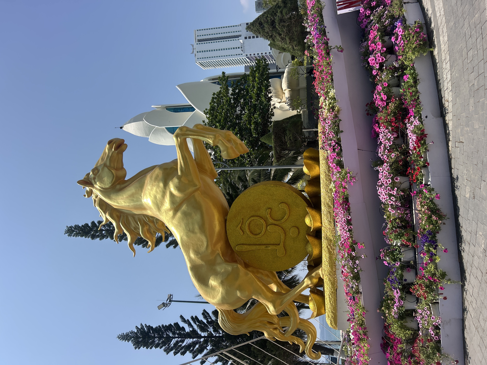 The image shows a golden horse statue rearing up on its hind legs next to a circular golden plaque with the number "25" on it. Behind the statue are trees, a white futuristic building, and a tall white building with many windows. To the right of the statue is a vertical arrangement of flowers. The statue appears to be located on a paved surface. Based on the buildings in the background, the location is likely the city of Nha Trang, Vietnam.