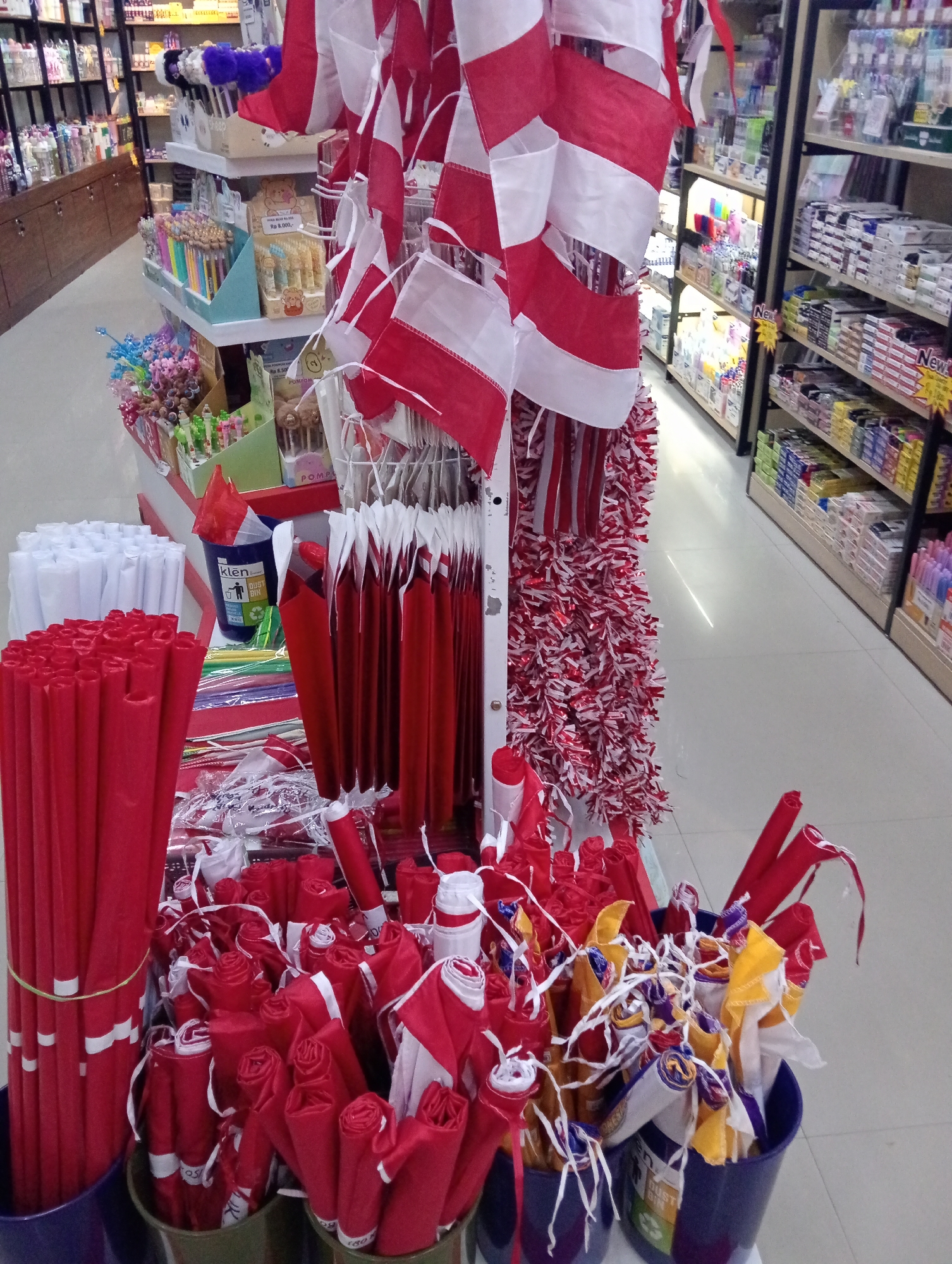The image depicts a retail display of patriotic merchandise, primarily in red and white, set up within a store aisle. The merchandise includes small Indonesian flags attached to sticks, red and white decorative streamers, and bundled red paper rolls. In the foreground, several containers hold these items. In the background, shelves lined with various products, including stationery and other retail goods, are visible. The store appears to be a general store or a shop selling party supplies, and based on the patriotic theme and the specific merchandise, it is likely located in Indonesia, possibly in a city like Pringsewu, where such displays are common during national holidays. There are no people visible in the image.