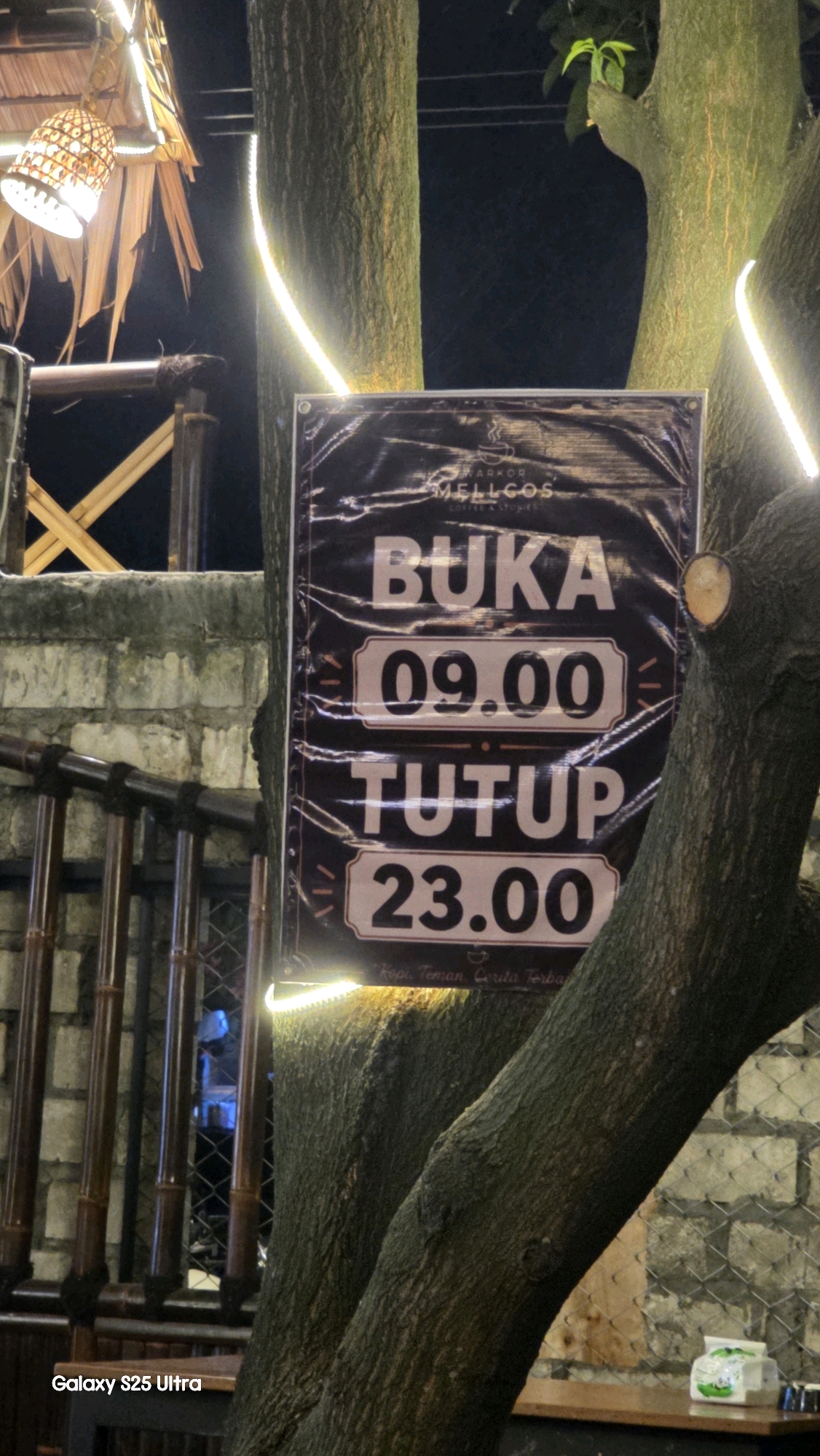 The image captures a sign indicating the operating hours of a business, likely a coffee shop or eatery, attached to a tree trunk in Juwana, Indonesia. The sign, written in Indonesian, displays "BUKA" (Open) above the time "09.00" and "TUTUP" (Close) above "23.00". The background reveals a rustic outdoor setting with bamboo railings, brick walls, and warm lighting from decorative lamps. The scene is illuminated by string lights wrapped around the trees, creating a cozy ambiance. The sign itself appears to be made of dark material with bold, light-colored text and is slightly crinkled, suggesting it is a banner or poster. The lower portion of the image includes a watermark that reads "Galaxy S25 Ultra."
