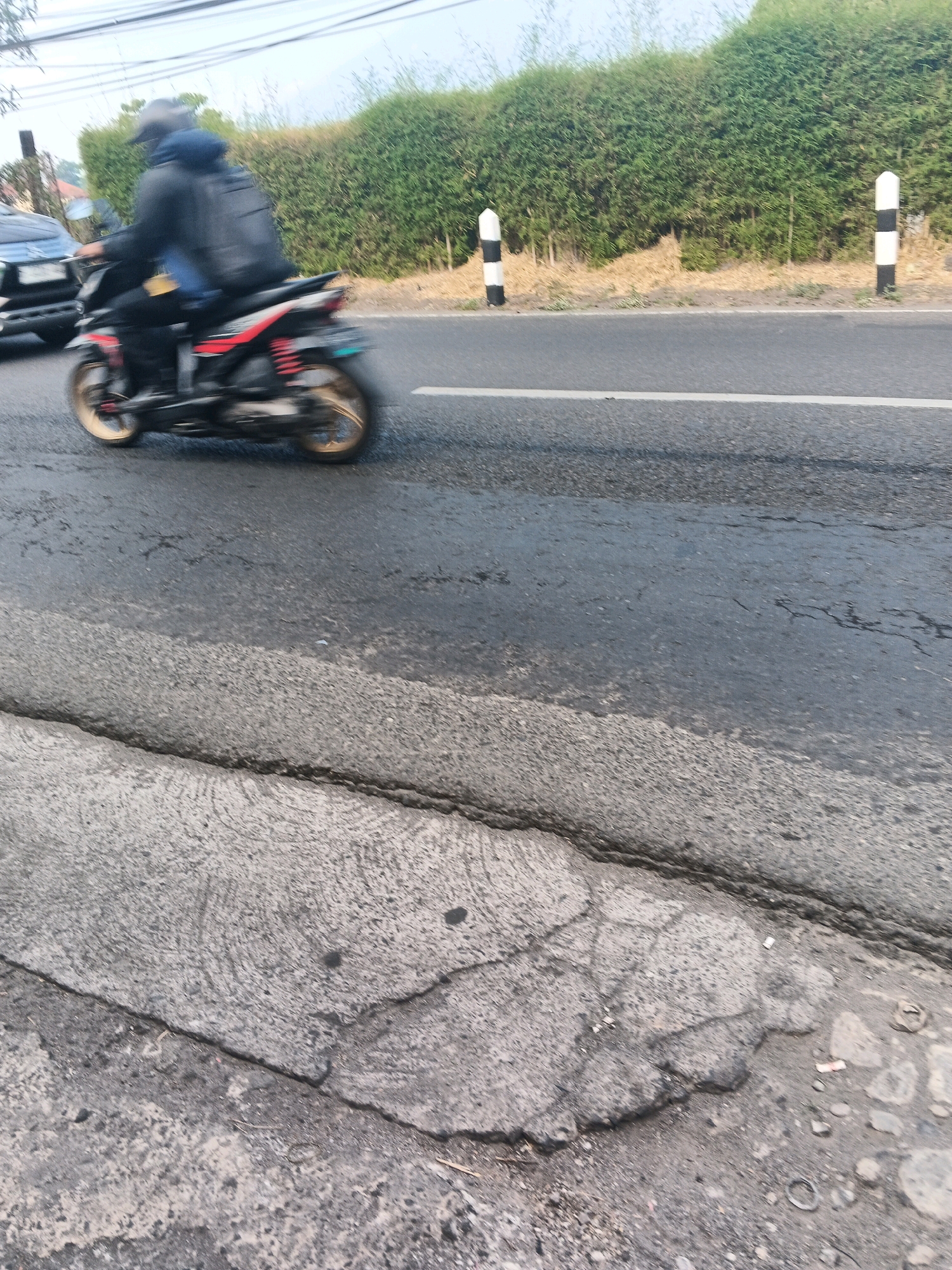 A person rides a motorcycle on a road in Ungaran, Indonesia. The person is wearing a dark jacket, blue jeans, and a helmet. They have a backpack on their back. The motorcycle is black with red accents and gold wheels. The road is paved and appears to be in good condition, with a white line dividing the lanes. A dense green hedge lines the side of the road, and there are two black and white striped bollards visible. The sky is overcast. The foreground shows a cracked concrete sidewalk. The image is taken from a low angle, and the motorcycle is in motion, creating a blur.