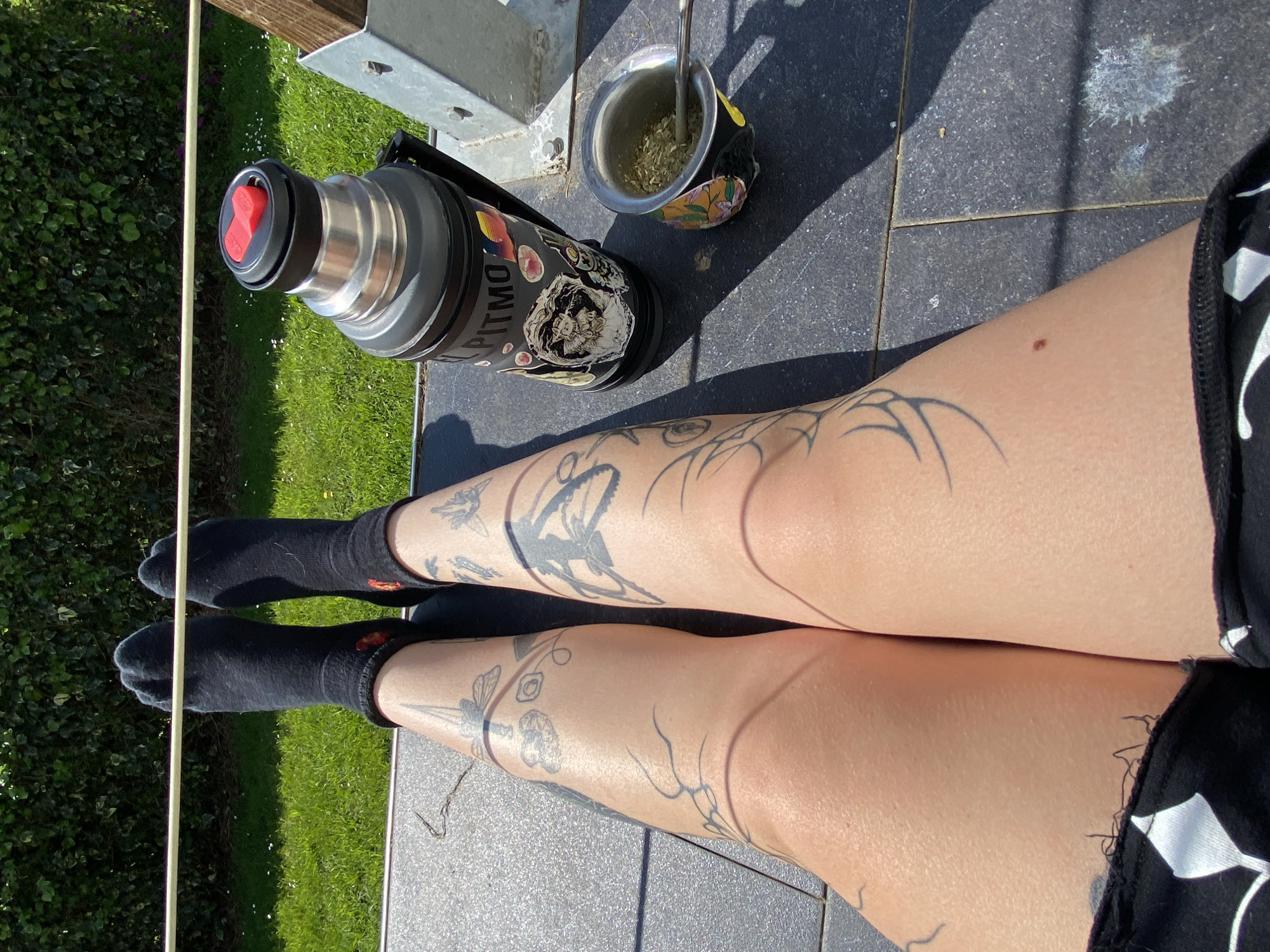 The image shows a person sitting with legs outstretched on a grey tiled surface, likely an outdoor patio. The person is wearing black socks and patterned black and white shorts. Both legs have multiple tattoos. A thermos with stickers, a mate cup with a metal straw, and a wooden pole are on the patio surface near the person. The background includes green grass and foliage. Based on the location, it is highly unlikely to be Villaviciosa, Spain as the question suggests. There is not enough information in the image to ascertain if the subject is in Villaviciosa, Spain, nor is there sufficient evidence to confirm its location.