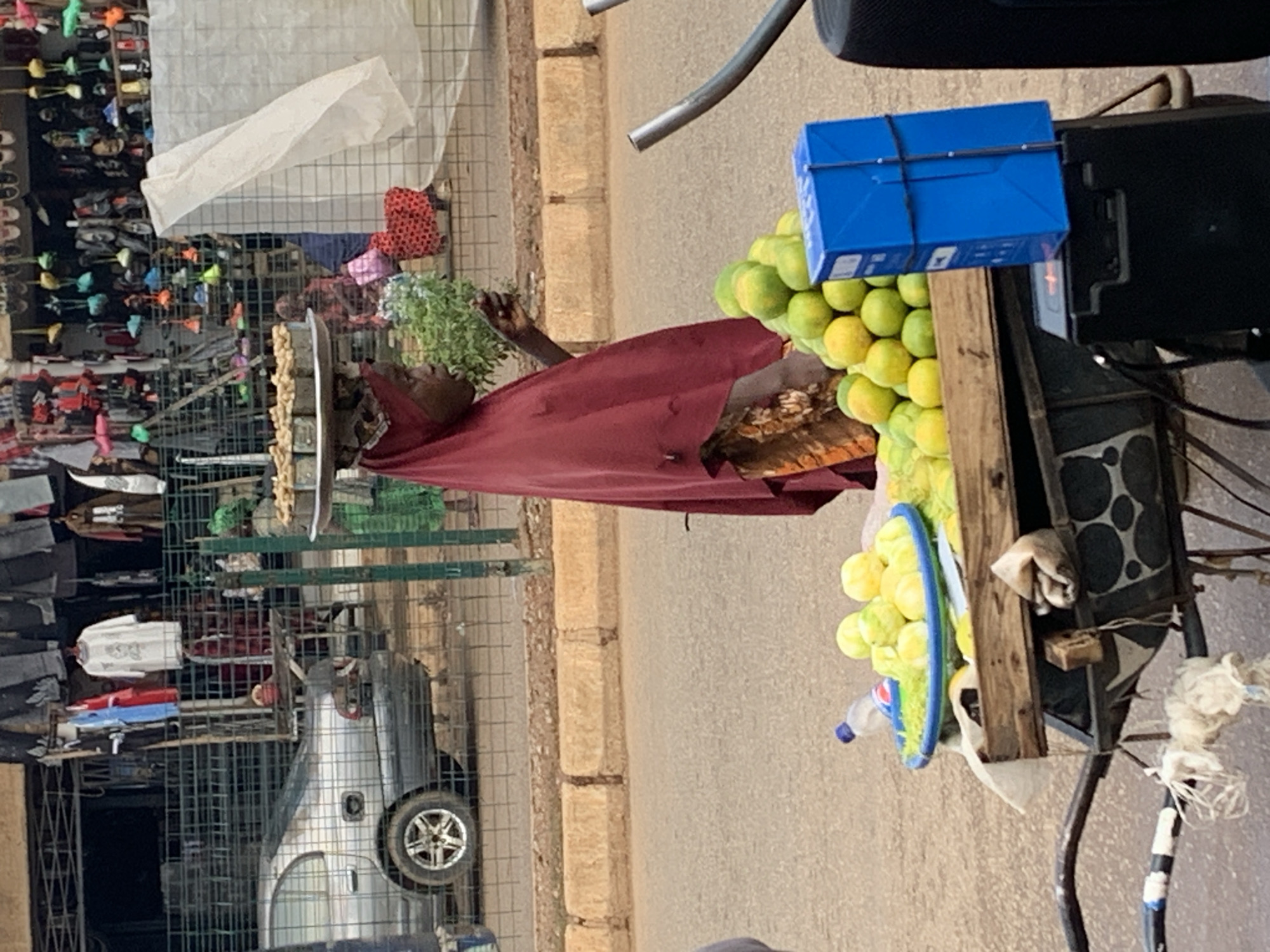The image depicts a street vendor in Bukuru, Nigeria, with a cart laden with goods. The primary subject is a person, likely the vendor, wearing a maroon garment and partially obscured by the cart. The cart is filled with a variety of items. Prominently displayed are stacks of yellow-green fruits, possibly citrus, arranged in a blue plastic basin. A blue box, secured with black straps, is attached to the side of the cart.
Behind the vendor, a rack filled with clothing and other items is visible, suggesting a market stall or a similar retail setup. A silver vehicle is parked nearby, partially enclosed within a metal grid structure. The background shows a rough, textured surface, likely a road or pavement, and a wall with layered stone blocks, indicative of the local architecture.
The scene appears to be an outdoor setting during daylight, though the specific time of day and weather conditions are not explicitly discernible. The overall impression is one of a typical street market environment in Nigeria. There is no visible text in the image that provides additional context.