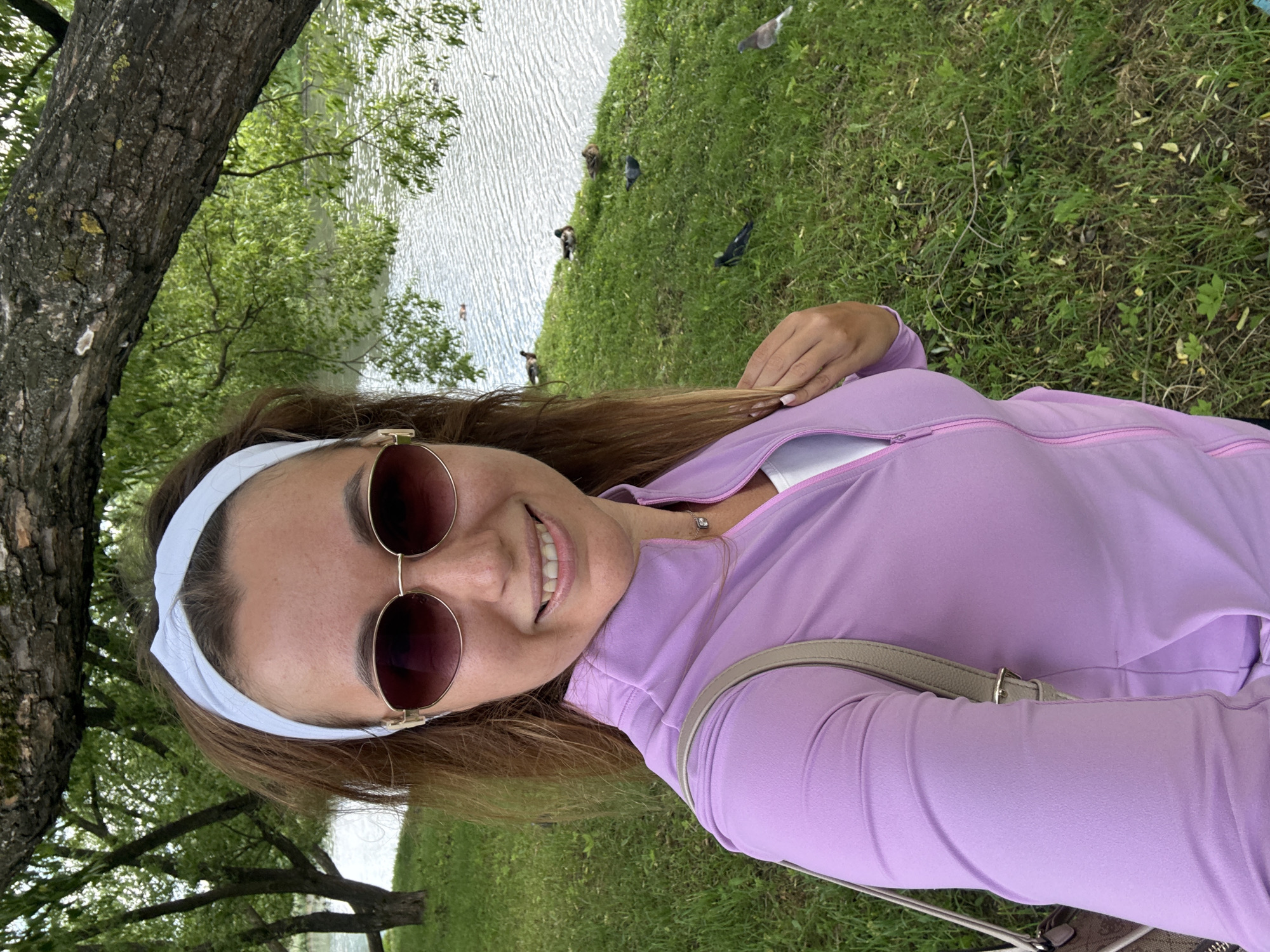 A woman is taking a selfie in a park. She is wearing a light purple zip-up jacket over a white top and a white headband. She has on sunglasses and is smiling. Behind her is a body of water with several ducks swimming, and a grassy bank with more ducks and pigeons. A large tree branch hangs over the scene from the left. The scene appears to be outdoors during the day.