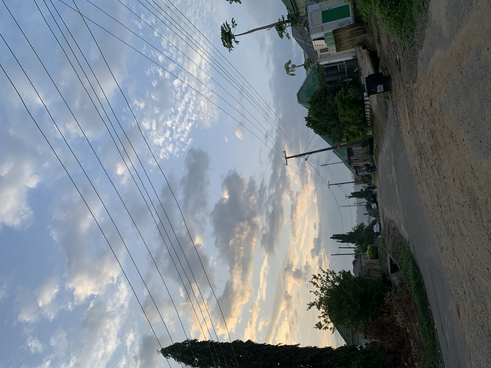 The image shows a street scene in Abuja, Nigeria, photographed from an unusual angle. The road, made of gravel and dirt, occupies the right side of the frame. Houses with green roofs and vegetation are visible on the right side of the road, while the left side displays a sky filled with clouds and several telephone wires. Tall trees are scattered among the houses. The sky shows a mix of white and gray clouds, and hints of sunlight, suggesting it may be early morning or late afternoon. In the distance, there appear to be parked vehicles on the road. There are no people visible in the scene.