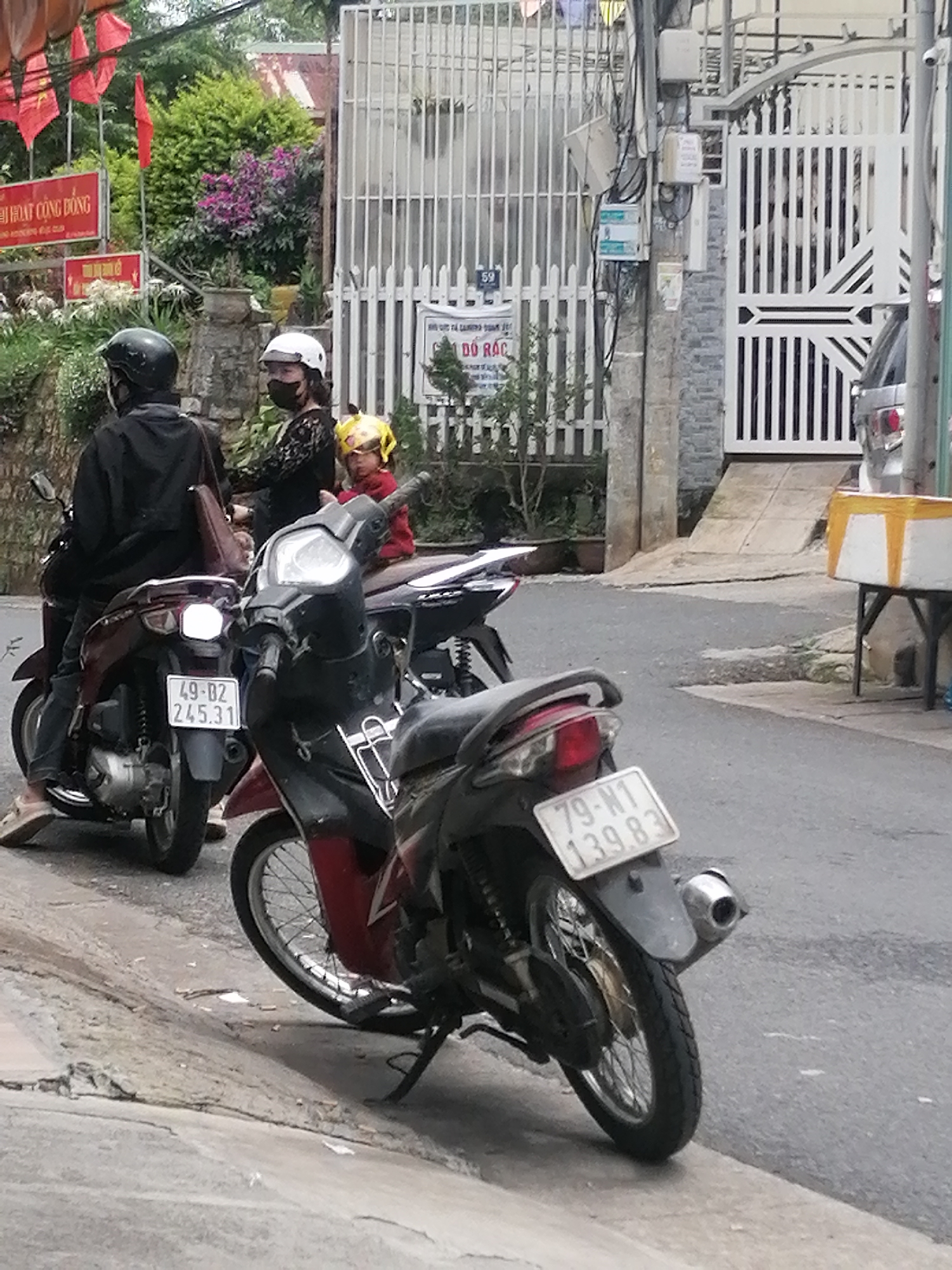The image shows a street scene in Da Lat, Vietnam. Two parked motorbikes occupy the foreground; the closer one has a license plate reading "79-N1 139.83". Behind it, a second motorbike has a license plate reading "49-B2 245.31".
Three people are seated on another motorbike behind the parked ones. The driver wears a black helmet and a dark jacket. A woman, wearing a white helmet and face mask, sits behind them, and a child with a yellow helmet sits behind her.
In the background, a white metal fence and gate can be seen. A sign with Vietnamese writing and red flags are visible on the left. The sign on the white fence reads "59" and "DO RAC". Power lines and an electrical meter are attached to a utility pole near the gate. A silver car is partially visible behind the gate. A table with a box on it sits near the gate. The road slopes uphill slightly.