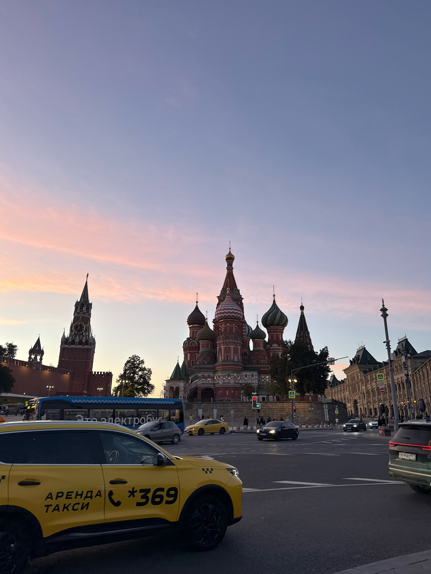 The image depicts a street scene in Moscow, Russia, with St. Basil's Cathedral and the Spasskaya Tower of the Moscow Kremlin visible in the background. The sky displays a gradient of pale blue to pink, indicating dusk or dawn. In the foreground, a yellow taxi with the inscription "АРЕНДА ТАКСИ" and the number "369" is prominently featured. Several other vehicles, including a blue bus and various cars, are on the road, suggesting a busy urban environment. Pedestrians are also visible on the sidewalks. The architecture of the buildings, characteristic of Moscow, and the presence of iconic landmarks confirm the location.