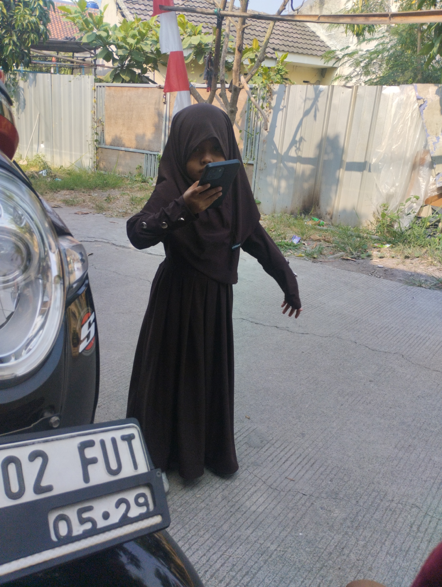 A young girl wearing a dark brown hijab and dress stands on a paved street, looking at a smartphone in her hands. To her left, a portion of a black car's front bumper and headlight are visible. The car has a license plate that reads "02 FUT 05.29". In the background, a corrugated metal fence, a wooden gate, and vegetation line the street. A red and white flag is also visible. The scene appears to be in Karangraharja, Indonesia, as indicated by the license plate format and general environment.