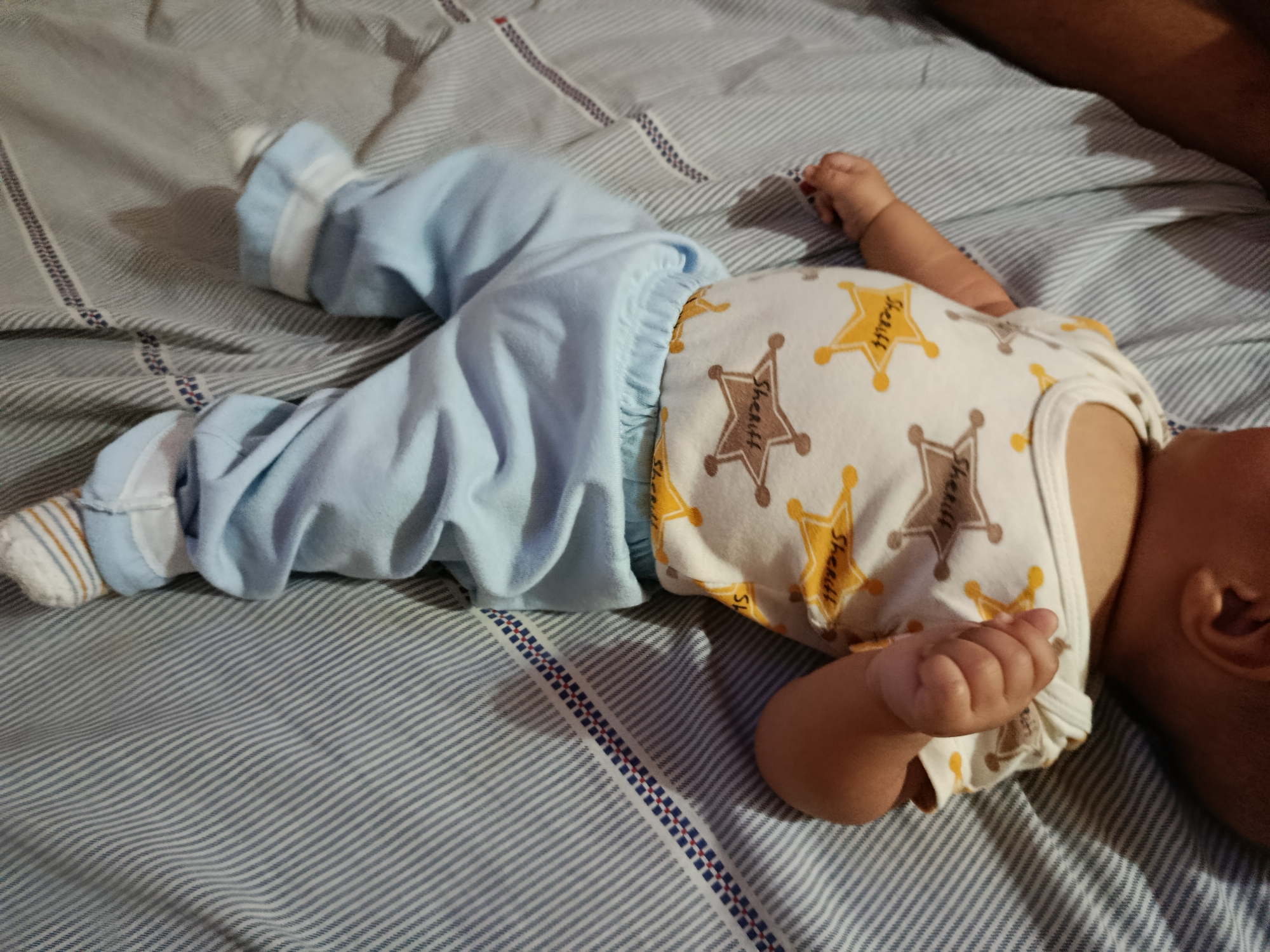 A newborn baby is lying on a striped bed in Altagracia, Venezuela. The baby is wearing a light blue onesie and white socks with orange and blue stripes. The onesie has a sheriff badge pattern. The baby's right arm is extended and its hand is resting on its chest. The baby's head is turned to the right and its face is not visible. The bed is made of blue and white striped fabric.