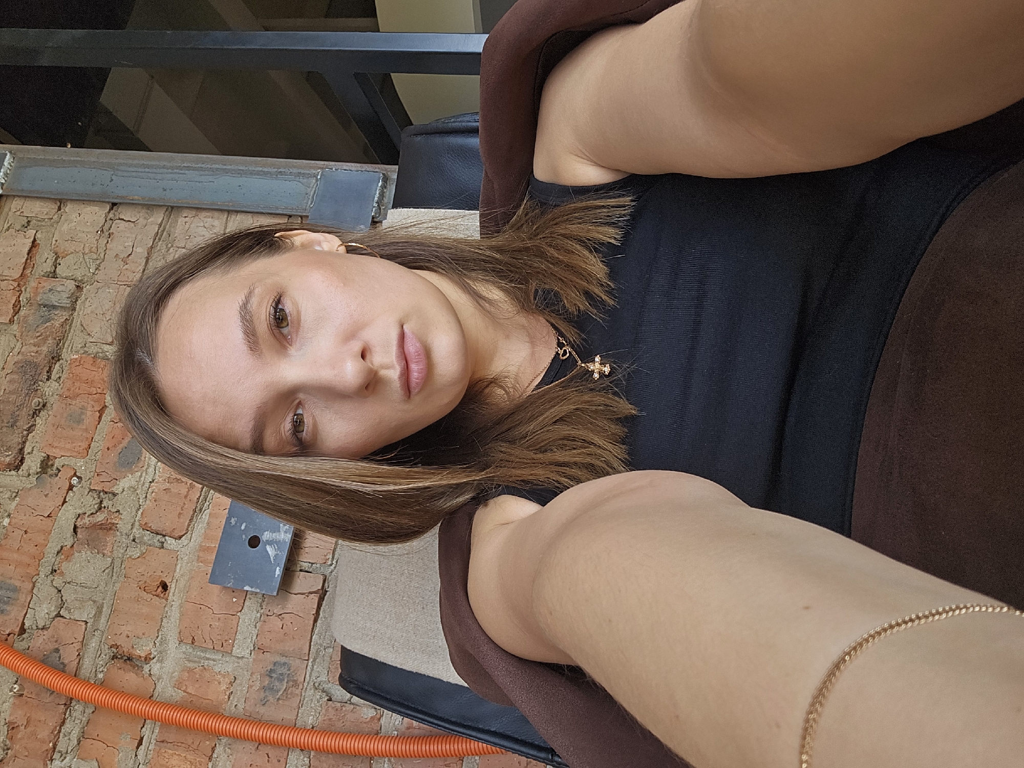 The image is a selfie of a woman with light brown hair, wearing a black top and gold jewelry. She appears to be sitting in a chair with brown upholstery. The background includes a brick wall, what looks like a metal bar, and a bright orange cable or pipe running along the base of the wall. The location in the city of Chaikovsky, Russia cannot be determined from the provided image.