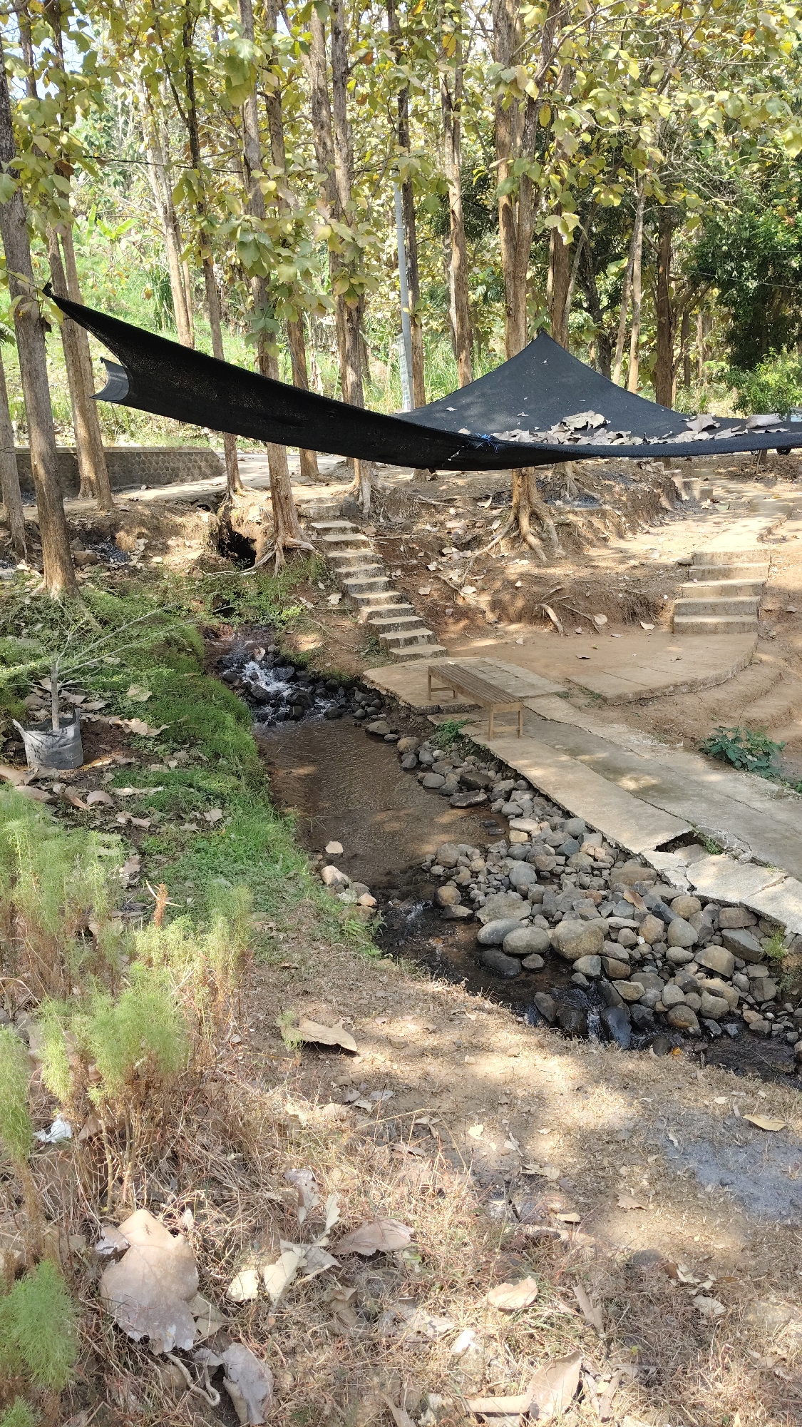 The image depicts a park area with a small stream flowing through it, set within a forest of deciduous trees. The scene is located in Kresek, Indonesia. In the foreground, dry grass and fallen leaves cover the ground. A narrow stream, with water flowing over rocks, meanders through the middle ground. On the right bank of the stream, a concrete pathway and a wooden bench are visible, with stone steps leading up to a higher level. On the left bank, more grass and a potted plant are present. Overhead, a black tarp stretches between trees, providing shade. The background consists of more trees and sunlight filtering through their leaves. The environment appears natural and somewhat rustic, with man-made elements integrated into the landscape. There are no people or animals visible in the image.