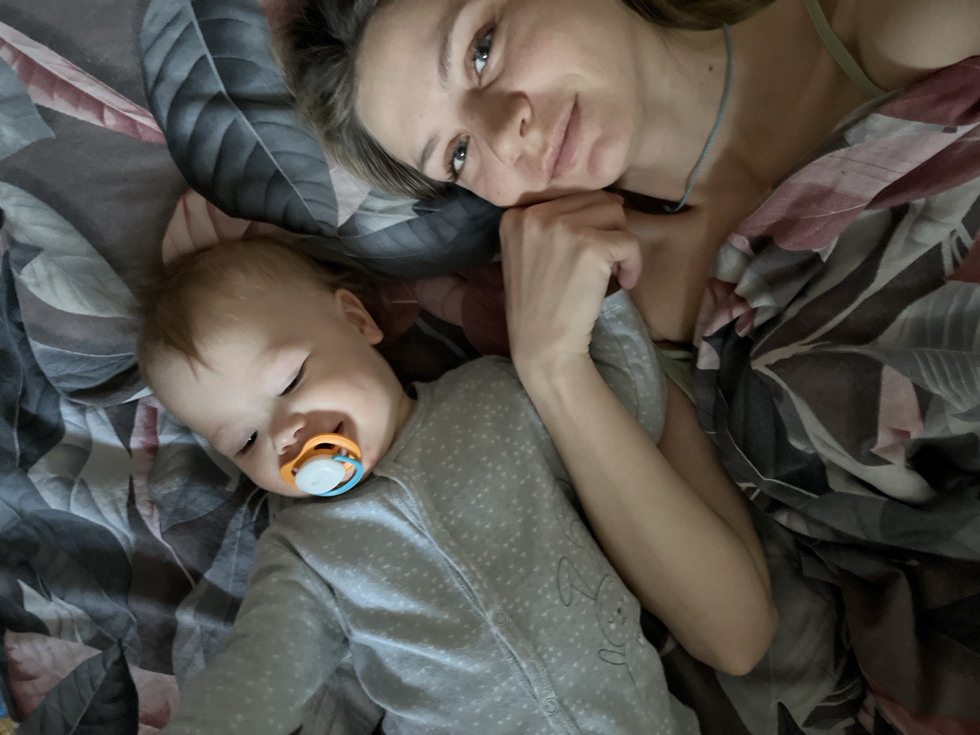 An indoor scene in Chaikovsky, Russia, depicts one adult female and one infant lying on a bed. The precise location within the city is not discernible from the image. The adult, positioned in the lower frame, lies on her back with her head towards the left, looking directly at the camera. She has light brown hair and a light blue string necklace visible on her neck, with a light-colored strap (possibly green) visible on her shoulder. The infant is positioned above the adult, lying on its back with its head near the adult's shoulder, facing towards the right side of the frame. The infant has light hair and a pacifier with an orange base, white nipple, and blue ring in its mouth, exhibiting a slight smile. The infant wears a light grey long-sleeved onesie with white polka dots and a grey cartoon bunny graphic on the chest. The adult's right hand is positioned near the infant's left arm, indicating proximity and interaction. Both individuals are resting on bedding that features a dark grey, white, and pink botanical pattern of large leaves.