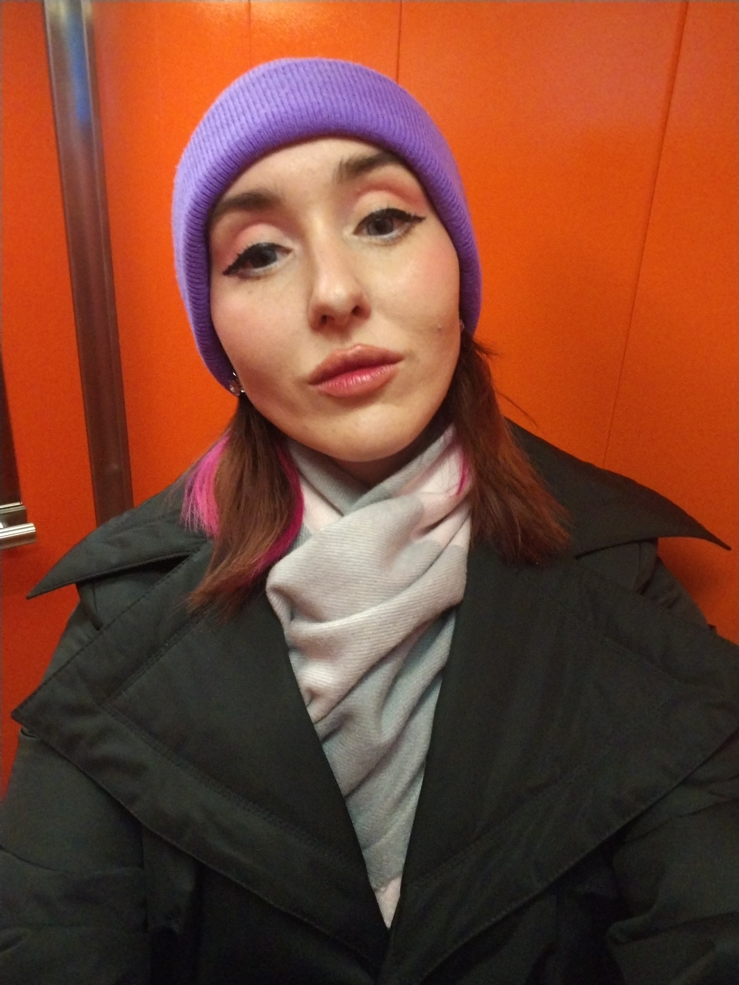 A woman wearing a purple beanie, a gray scarf, and a black coat is in an elevator with orange walls. The woman has pink highlights in her hair. A metal handrail is visible on the left side of the image. There is no evidence to support this image being taken in the city of Izhevsk, Russia.