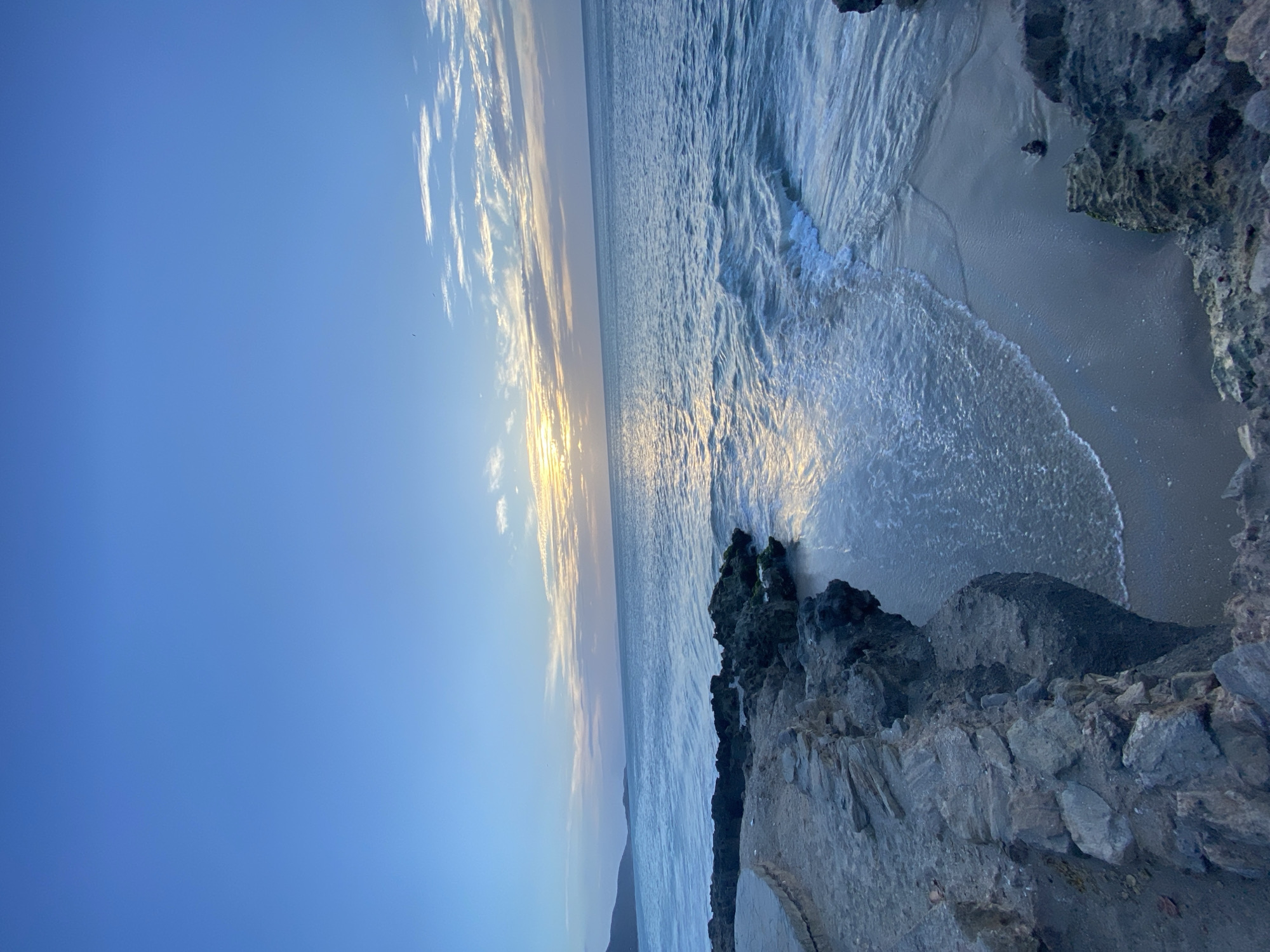The image shows a view of the ocean at sunset from a rocky coastline. The sky is blue, with streaks of clouds illuminated by the setting sun. The ocean water is light blue, with waves crashing onto a sandy beach. Large, grey rocks are in the foreground, forming a cliff or a rugged shoreline. The photo appears to be taken in Juan Griego, Venezuela.