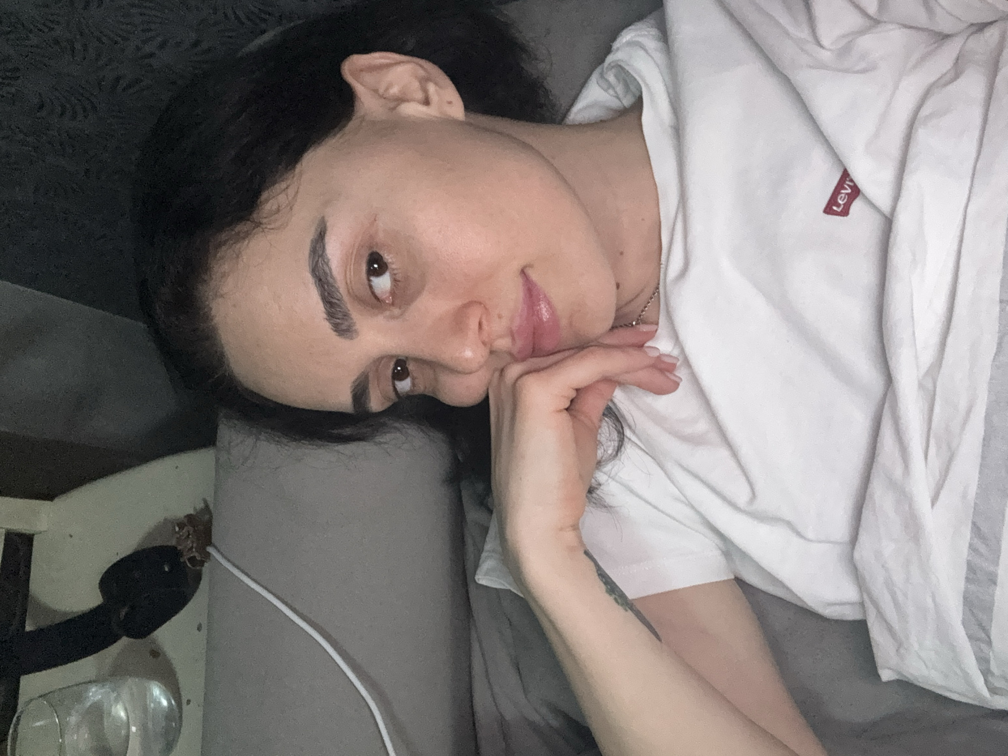 A woman is lying on a gray bed, wearing a white t-shirt with a red Levi's logo, and propping her head up with her hand. Her dark hair is loosely arranged around her face. An object with a black plug and a white cable are positioned on a nightstand next to her. The photo was most likely taken indoors. No specific information suggests this scene is located in Tbilisi, Georgia.