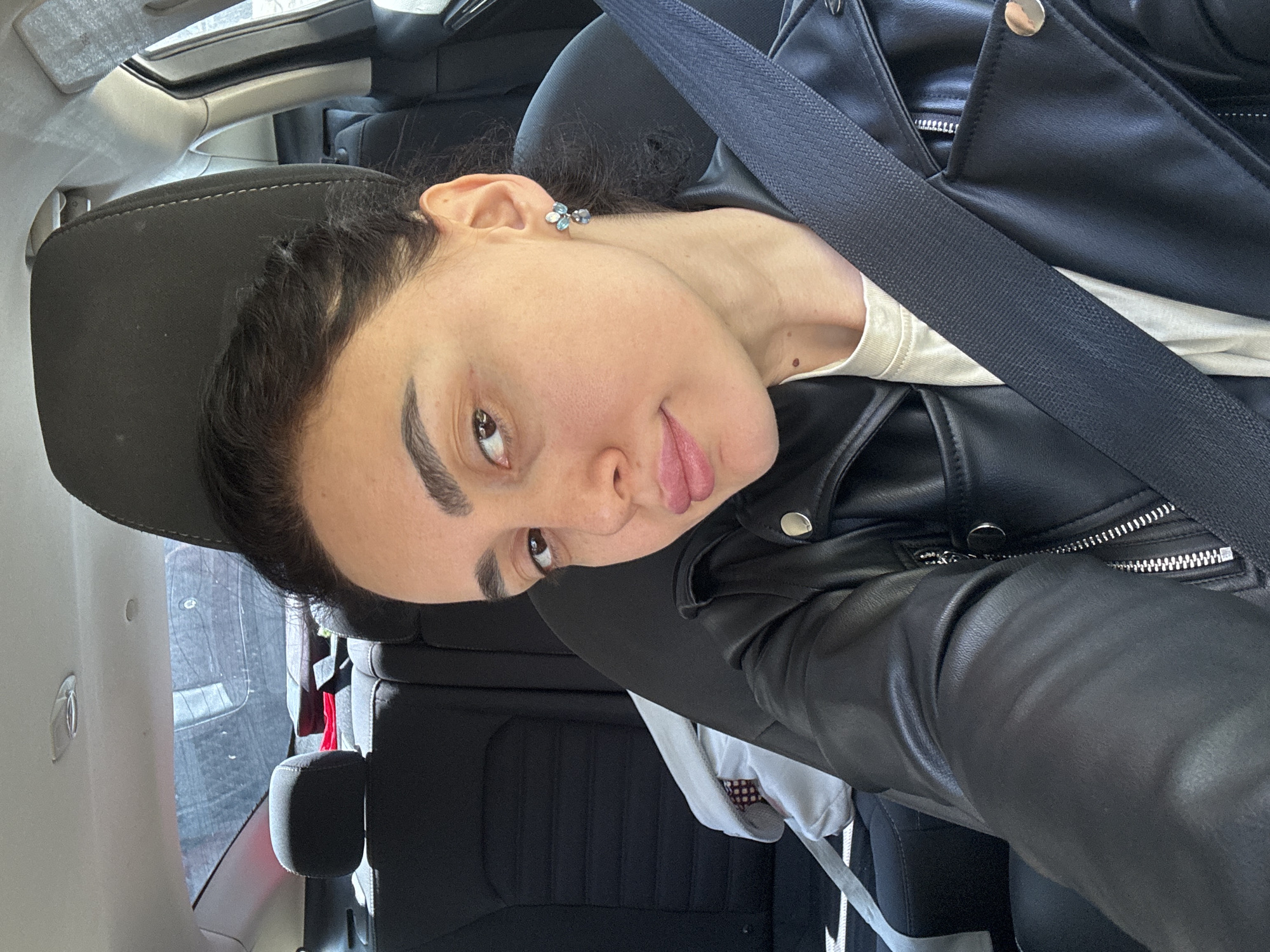 A young woman is in a car, wearing a black leather jacket, a white shirt and a seat belt. She is looking at the camera with an eyebrow raised and her tongue slightly sticking out. The car is likely moving, as suggested by the blurry background. The location is potentially Tbilisi, Georgia, although there is no direct evidence to confirm this.