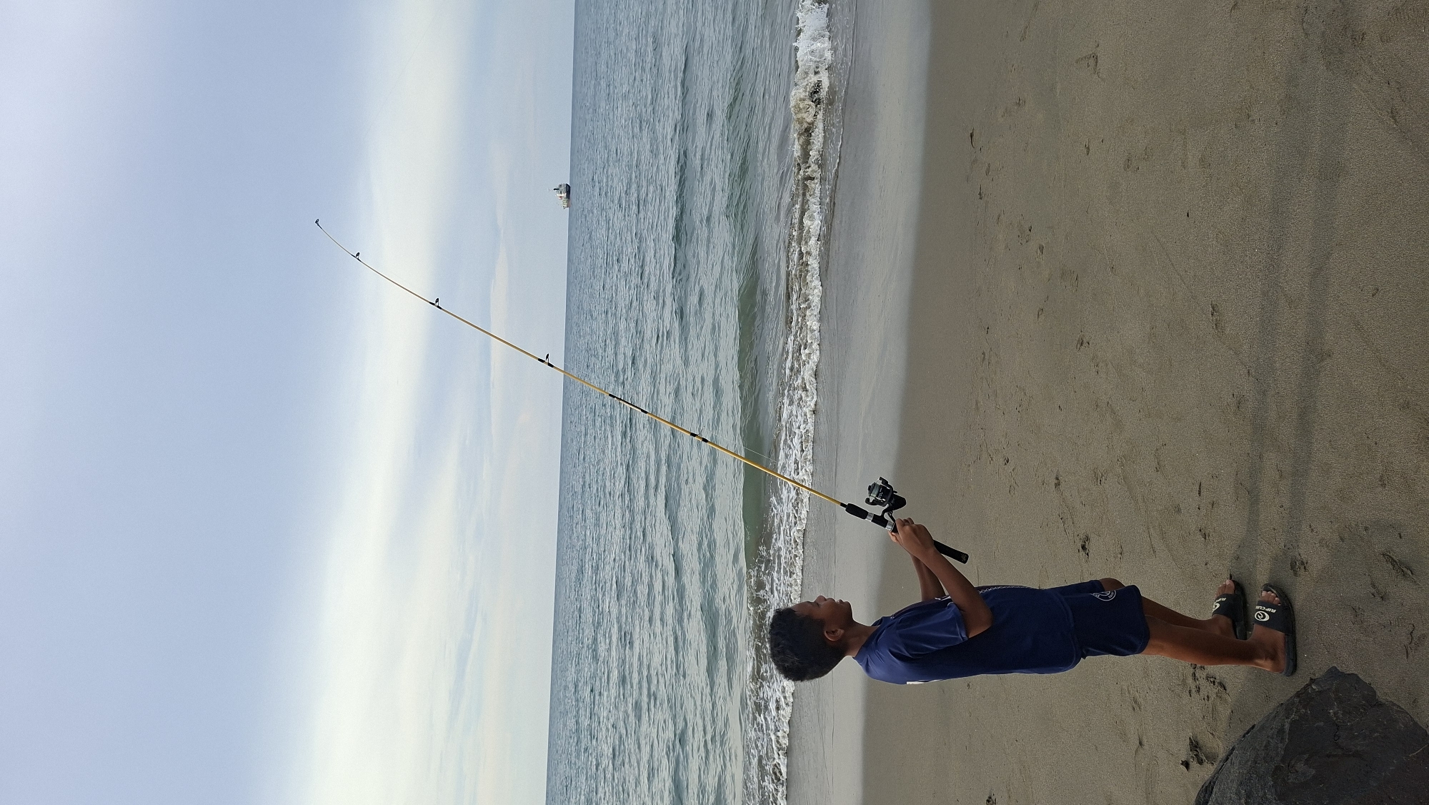 A young boy is fishing on a sandy beach in Lhokseumawe, Indonesia. He is wearing a blue t-shirt and blue shorts, and black flip-flops. He is holding a fishing rod with a reel, and is looking out at the ocean. The ocean is calm, with small waves rolling onto the shore. In the distance, a large ship is visible on the horizon. The sky is cloudy and overcast. The sand on the beach is light brown and has some footprints on it. There is a large rock on the left side of the image.