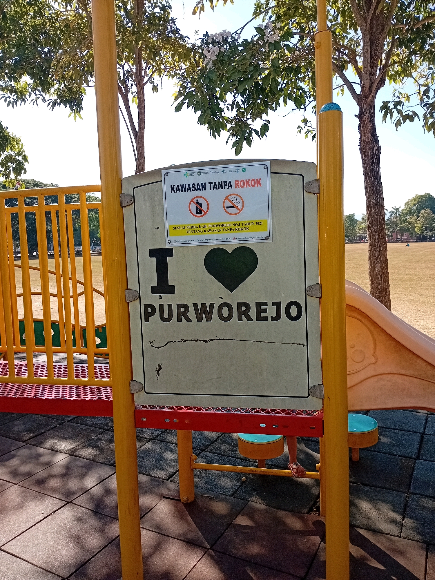 A "No Smoking" sign is affixed to a yellow playground structure in Purworejo, Indonesia. The sign, with a white background, displays the Indonesian text "KAWASAN TANPA ROKOK" which translates to "No Smoking Area". Below this, it states "SESUAI PERDA KAB. PURWOREJO NO. 4 TAHUN 2021 TENTANG KAWASAN TANPA ROKOK" which translates to "In accordance with Regional Regulation of Purworejo Regency Number 4 of 2021 concerning No Smoking Areas". The sign also features a stylized heart shape and the word "PURWOREJO" in large black letters. The playground equipment, primarily yellow and red, is set on a patterned rubberized surface. In the background, a grassy field with scattered trees and a few distant figures can be observed under a clear sky.
