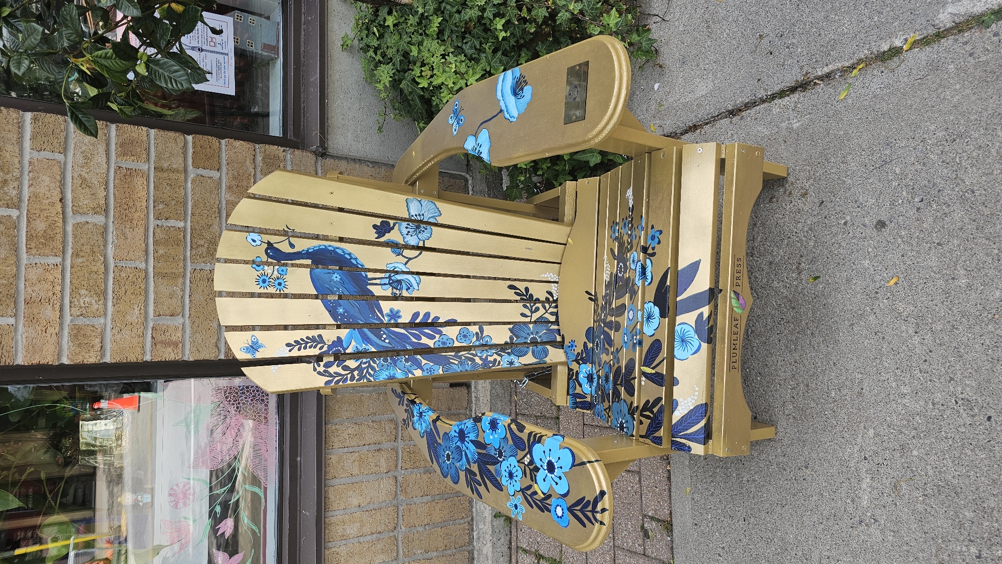 This image features a gold Adirondack chair painted with a vibrant blue floral and peacock design. The chair is placed on a concrete sidewalk next to a brick wall. To the left, a window of a building is visible, showing reflections of plants and possibly shelves with items. Above the chair, green foliage from a plant hangs down.
The chair's design includes a large peacock on the backrest and seat, surrounded by various blue flowers, leaves, and a butterfly. The flowers are detailed with dark blue outlines and lighter blue centers. The overall aesthetic is artistic and decorative.
On the side of the chair's leg, the text "PLUMLEAF PRESS" is visible vertically, suggesting it might be a custom-designed piece or part of a public art installation. The lighting suggests it is daytime, with diffused light, possibly indicating an overcast day or shade. No people are present in the image. The setting appears to be an outdoor area in front of a commercial building, possibly in Oakville, Canada, as indicated by the location context.