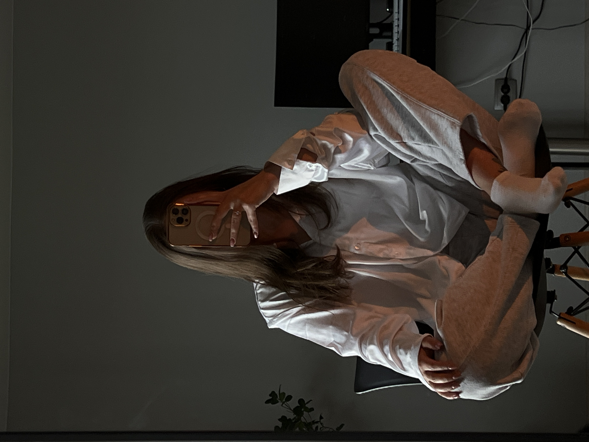 The image depicts an individual seated on a modern chair in an indoor setting, identified as a private room in the city of Ternopil, Ukraine. The subject, with long, straight dark brown hair, has their face completely obscured by a gold-colored smartphone held directly in front of them. They are wearing a loose-fitting white long-sleeved button-up shirt, light grey sweatpants, and white socks. Their legs are crossed at the ankles in a relaxed posture, with knees elevated. The subject's visible left hand, holding the phone, shows light pink or nude nail polish and a ring on the ring finger.
The room is dimly lit, with a prominent light source originating from the right side of the frame, illuminating the subject's right arm, shirt, and parts of their legs, creating strong highlights and shadows. The background features a plain, dark grey wall. To the left of the subject, a dark-leaved plant is partially visible. To the right, a dark computer monitor, a keyboard, an electrical outlet, and multiple cables are arranged on a surface, likely a desk. The chair has a dark seat and light-colored wooden legs with black metal cross-bracing. The overall scene conveys a casual, private, and somewhat introspective atmosphere.