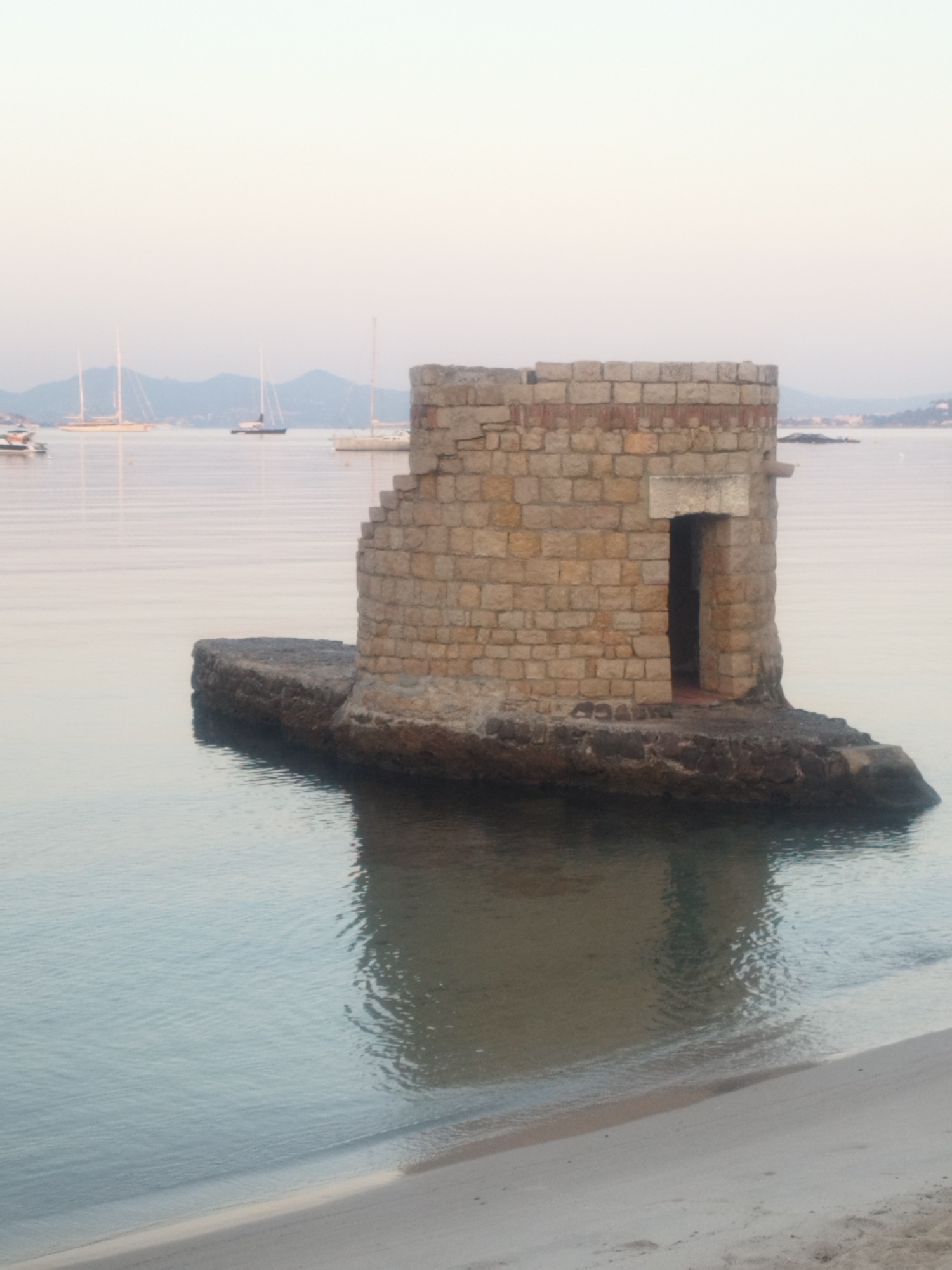 The image depicts a small, stone tower built on a rocky outcrop in the water, near a sandy beach. The tower is constructed from light-colored, rough-hewn stones, with a dark, recessed doorway. The water is calm and reflects the sky and the tower. In the background, several sailboats are visible on the sea, with a hazy coastline and hills in the distance. The sky is pale and overcast, suggesting early morning or late evening. This scene is located in Antibes, France, and is likely a historical watchtower or a small fortification. There are no people visible in the image. The environment is tranquil and maritime.