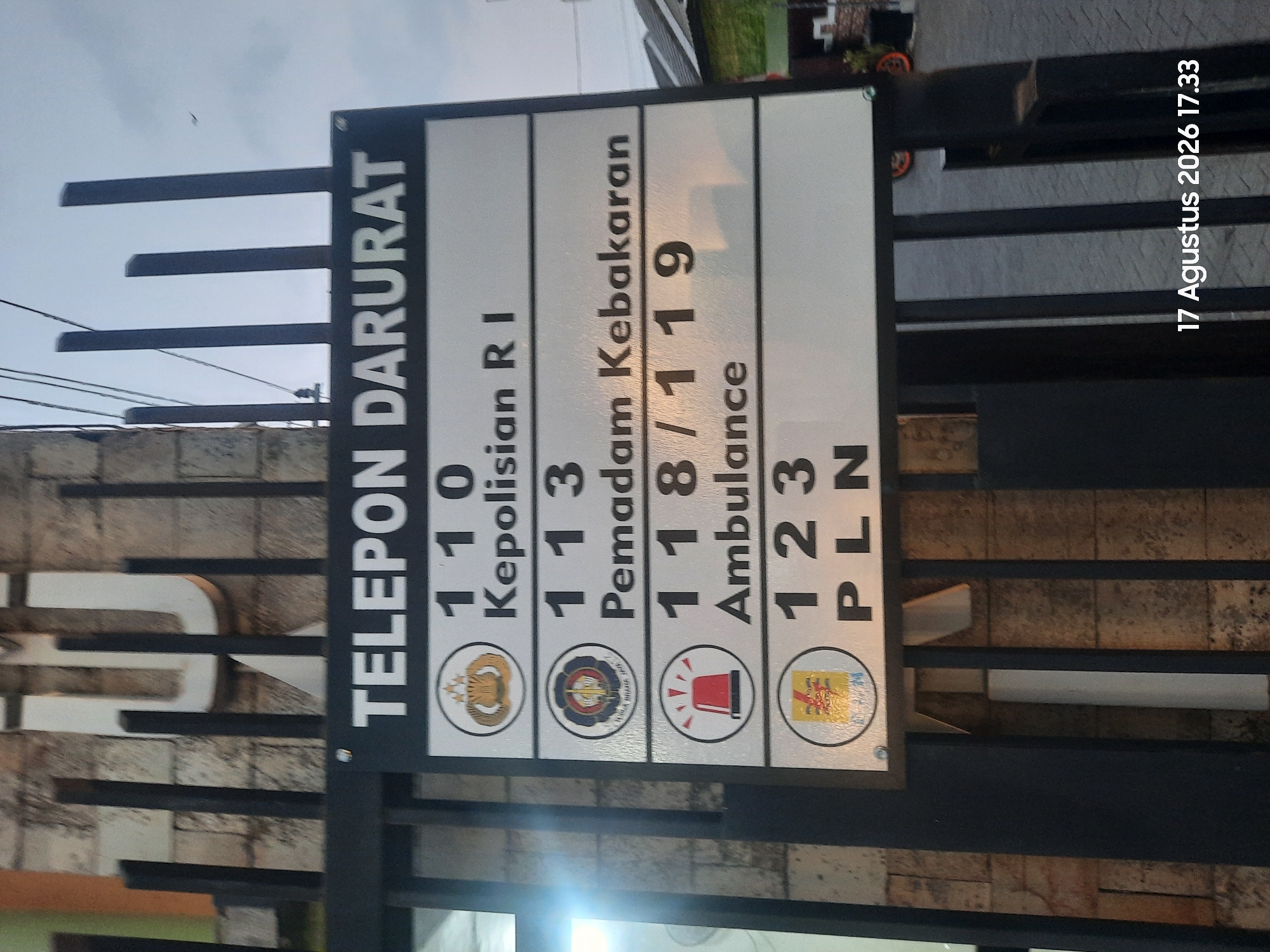 A sign board displaying emergency telephone numbers is mounted on a black metal gate. The sign has a dark blue header with white text that reads "TELEPON DARURAT" (Emergency Telephone). Below the header, there are four sections, each with a number and a description of the service. The first section shows the number "110" and the text "Kepolisian R I" (Indonesian National Police). The second section shows "113" and "Pemadam Kebakaran" (Fire Department). The third section displays "118 / 119" and "Ambulance". The fourth section shows "123" and "PLN" (State Electricity Company). Each section, except for the last one, has a small circular logo or icon next to the number. The logos are for the Indonesian National Police, the Fire Department, and an ambulance siren. The gate is made of black vertical bars and is positioned in front of a brick wall. The background shows some buildings and a cloudy sky. A timestamp "17 Agustus 2026 17.33" is visible in the bottom right corner of the image.