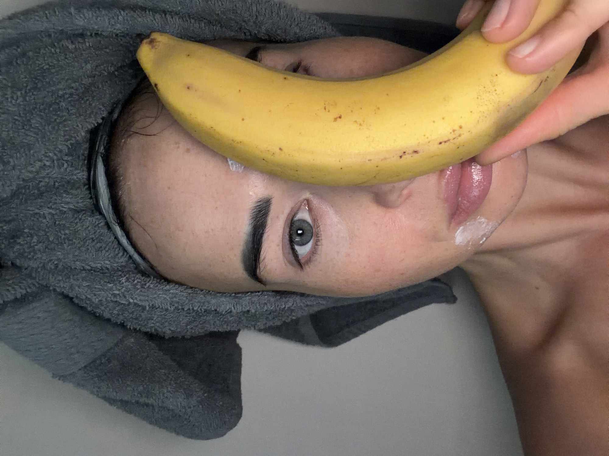 A close-up, top-down photograph captures a person, likely female, engaged in a beauty or skincare routine in an indoor setting. The individual is lying down, with their head wrapped in a fluffy grey towel. A ripe yellow banana is positioned horizontally across their face, extending from just above the eyebrows down to the chin, held in place by a hand visible near their mouth.
The person's left eye, a striking grey-blue, is open and looking directly at the camera. Their dark, well-defined eyebrows are prominent. White creamy residue is visible on their chin and lower lip, and a small smudge is near the inner corner of their left eye, suggesting the application of a facial product. Their lips are slightly parted, appearing glossy or moisturized. The right side of their face is largely obscured by the banana. The overall mood conveyed is calm and relaxed, possibly playful due to the banana's unusual placement. The background is a plain, light color. There is no visible text in the image. This scene is set in Saint Petersburg, Russia.