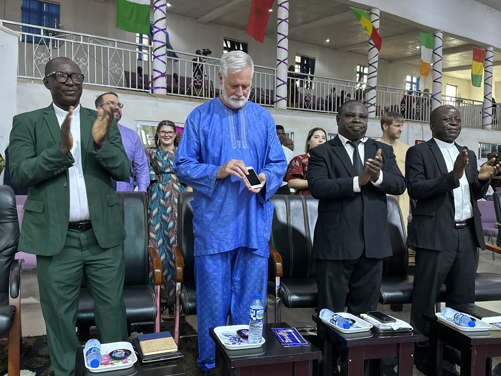 This is a medium shot, eye-level, outdoor photograph taken in the city of Yelwa, Nigeria. In the foreground, three men are standing. The man on the left is wearing a green suit and white shirt, glasses, and is clapping. The man in the center is wearing a blue traditional Nigerian outfit and is looking down at a white cell phone in his hand. The man on the right is wearing a black suit and white shirt, and is also clapping. Behind them, several people are seated in chairs, and in the background, flags and decorations are visible. The setting appears to be a public gathering or ceremony.
