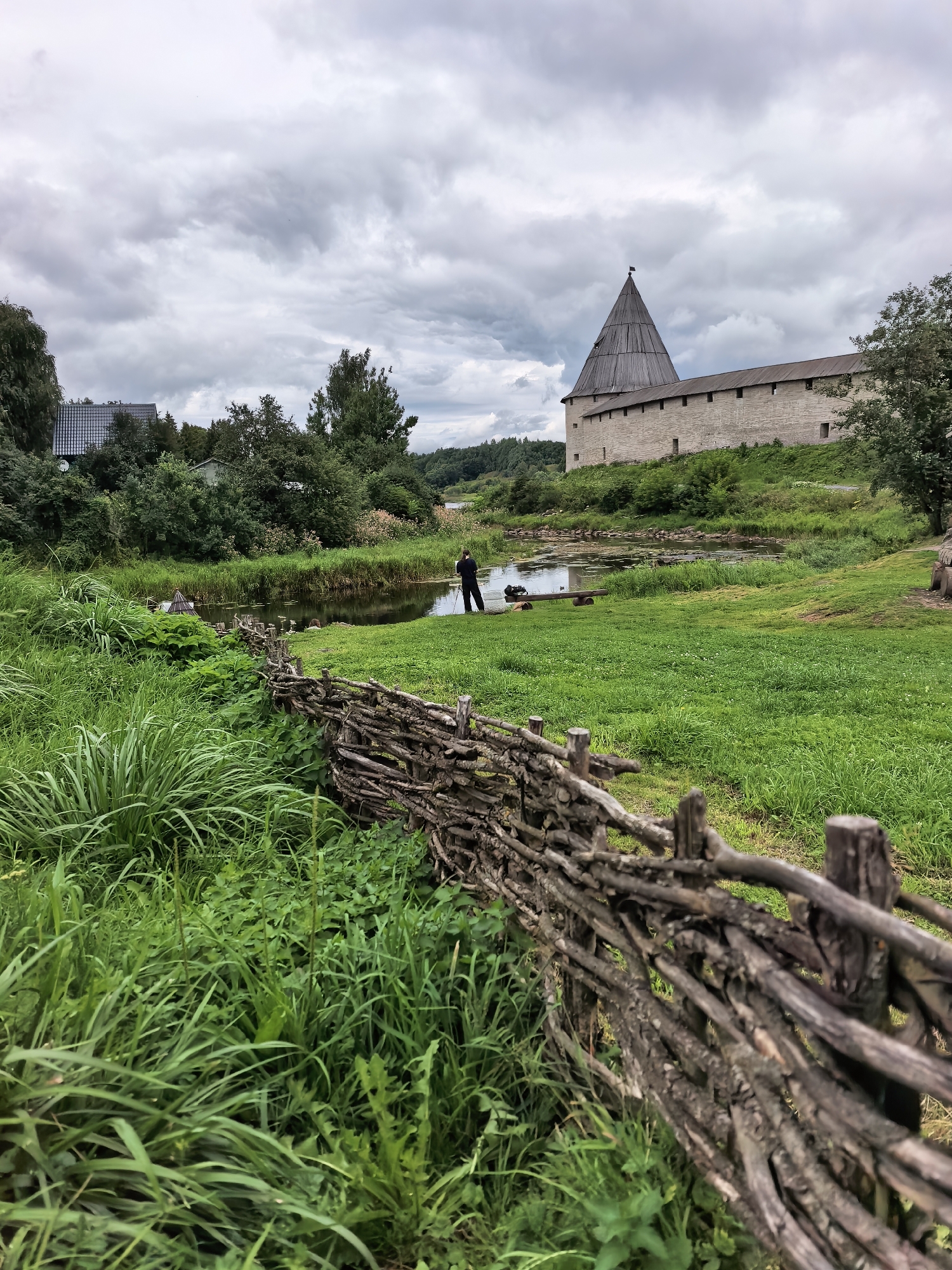 A person stands near a body of water in Staraya Ladoga, Russia, beside a rustic wooden fence. The scene depicts a historical fortification, likely the Staraya Ladoga Fortress, characterized by its stone walls and a conical wooden tower. Lush green grass covers the landscape, with trees and foliage surrounding the water and buildings. The sky is overcast with heavy clouds, suggesting a potentially cool or rainy day. The foreground is dominated by the textured, weathered wood of the fence, while the background features the imposing fortress architecture.