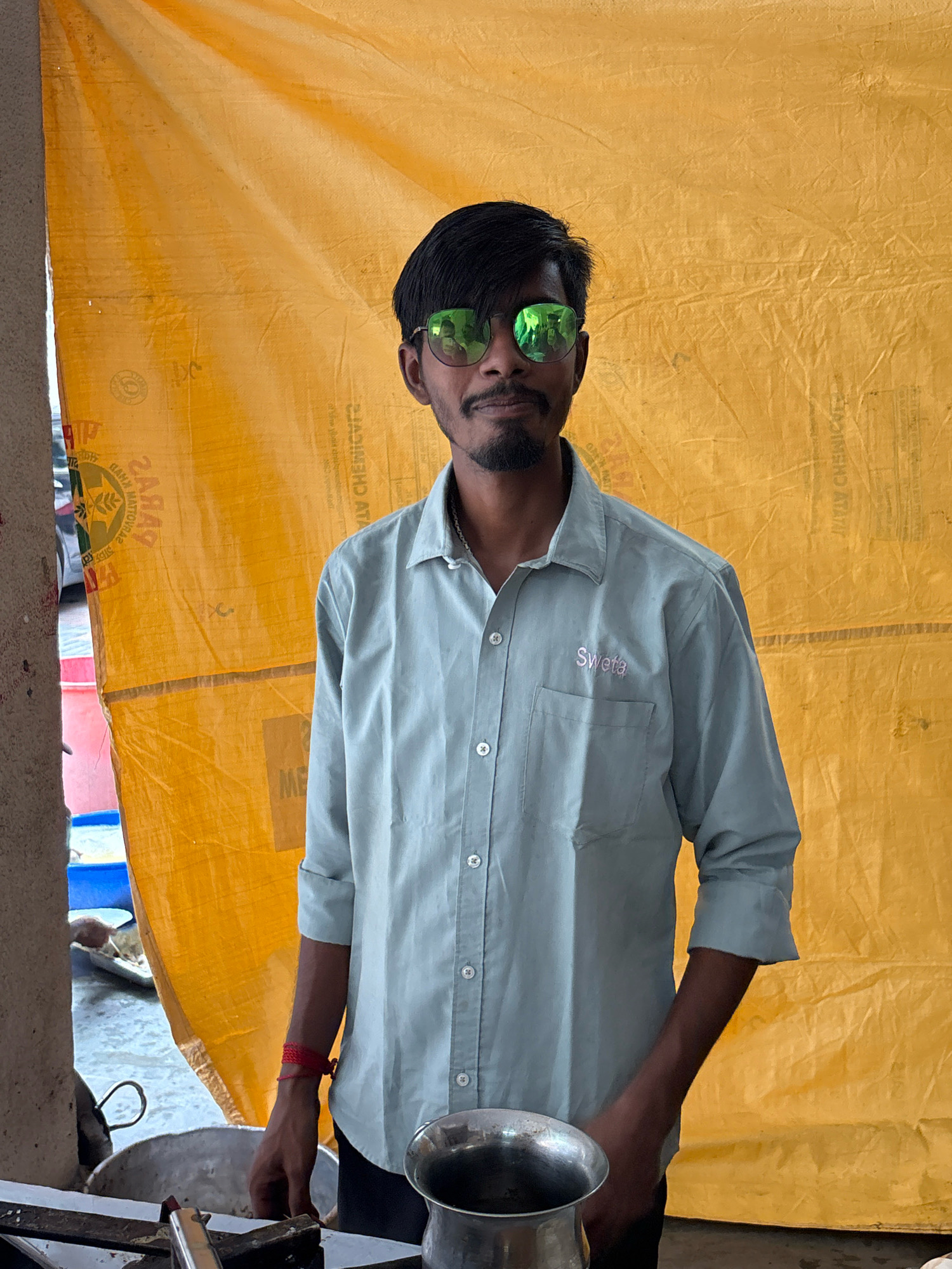A man stands in front of a yellow tarp in Howrah, India. He is wearing a light blue button-down shirt with a pocket on the left side, sunglasses with green lenses, and a goatee. A red string is tied around his left wrist. To his right, a silver bowl is visible. The yellow tarp is wrinkled and has text and symbols printed on it. The background appears to be outdoors, with some blurry objects and structures visible on the left side of the image.