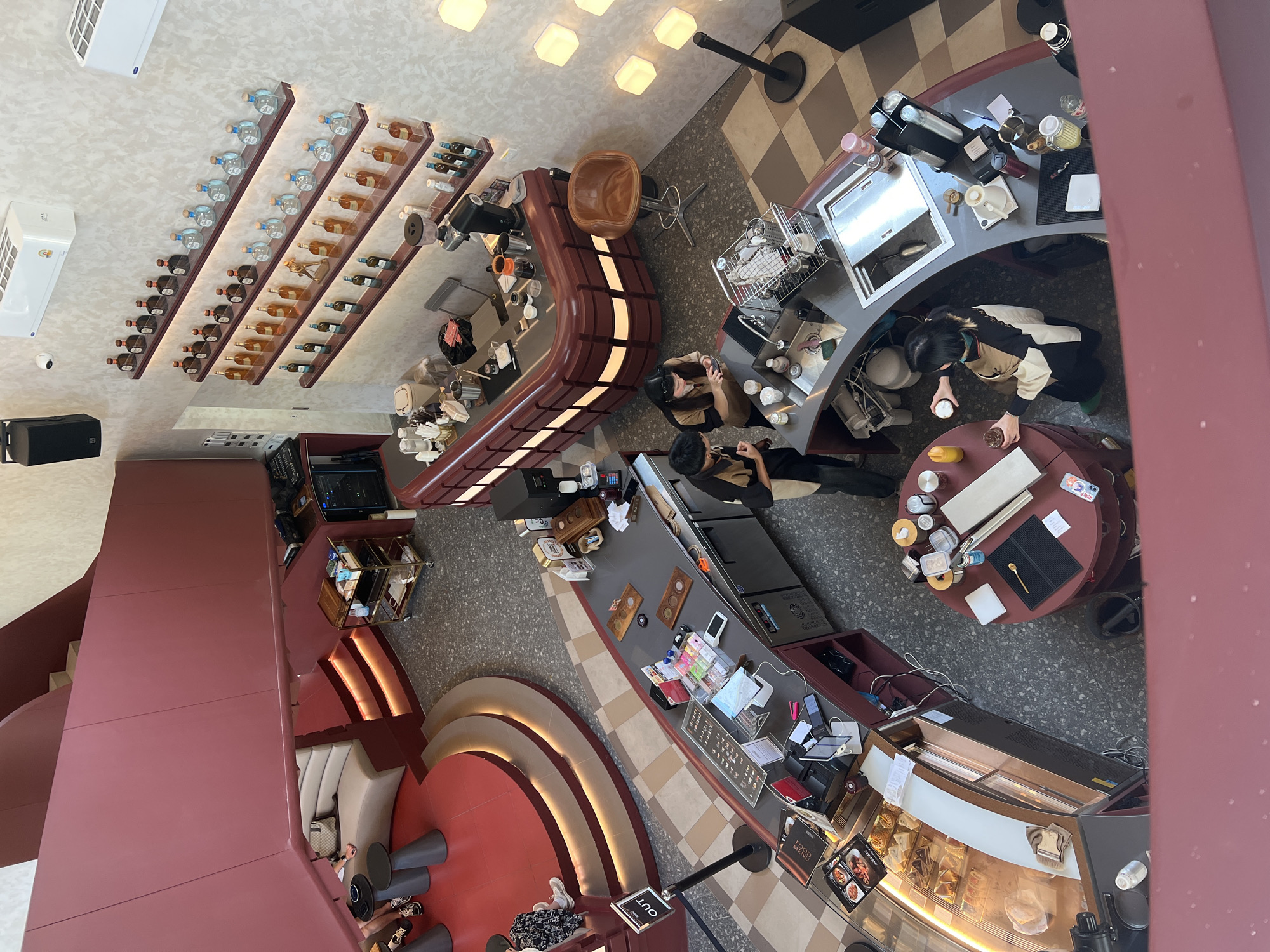 Captured from an elevated vantage point, the image shows the interior of a cafe or restaurant in Bangkok, Thailand. The architectural style incorporates curved elements and a color scheme dominated by shades of red and brown.
The cafe features several distinct areas:
* **Bar Area**: A curved bar occupies a significant portion of the space, equipped with coffee machines, sinks, and various containers. Employees are seen working behind the bar, interacting with customers.
* **Seating Area**: Rounded red booths with low couches are positioned along a wall, where patrons are seated.
* **Display Case**: A lit display case at the front showcases pastries and other food items.
* **Retail**: Shelves on the wall display bottles of liquor.
The cafe has a mix of checkered and speckled flooring, with contemporary lighting fixtures illuminating the space. Patrons are scattered around, engaging in activities such as ordering drinks, sitting at tables, and using electronic devices. Overall, the interior design blends modern aesthetics with elements of local Thai architecture, creating a unique ambiance within a bustling city environment.