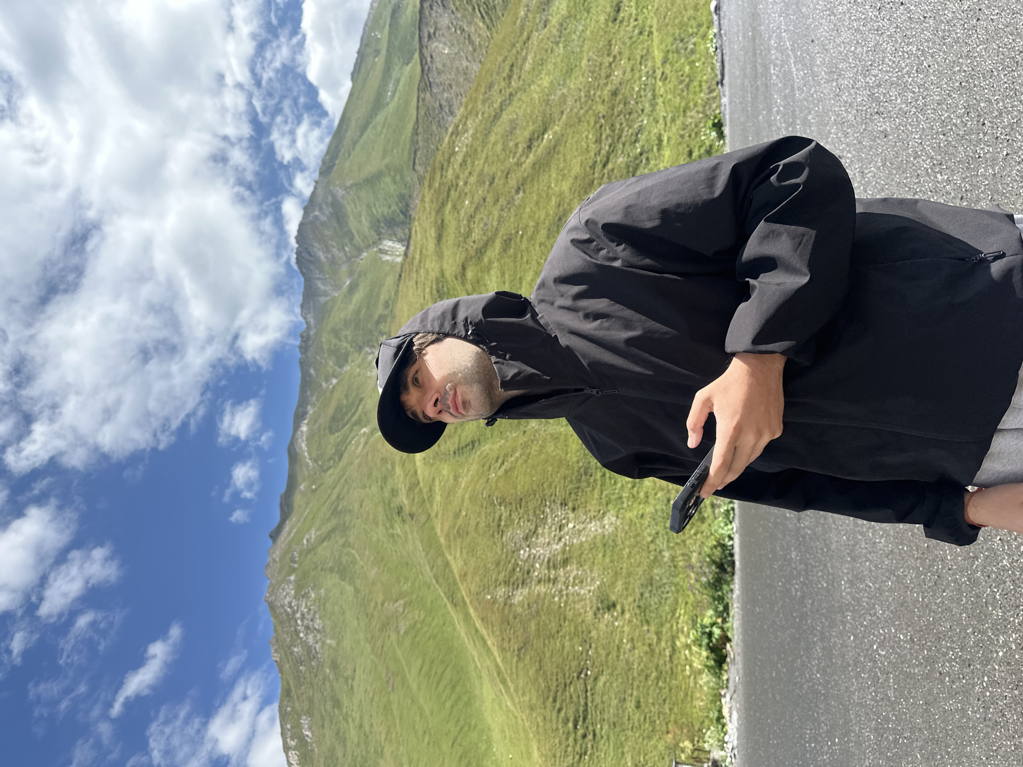A young man with a black baseball cap and hood up, wearing a black jacket and grey sweatpants, stands on a wet asphalt road holding a black smartphone. He gazes upwards and to the right. Behind him, lush green mountains under a blue sky with scattered white clouds rise up. The scene appears to be in Almasiani, Georgia, a region known for its mountainous terrain.