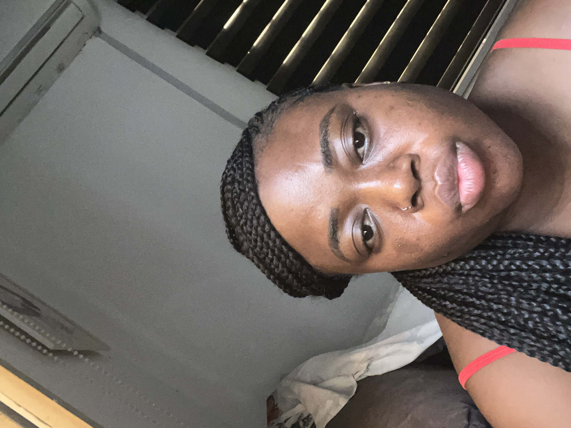 A person is facing the camera with a perspective from below. The person has dark skin, braided hair, and is wearing a red strap. The background includes white bedding and vertical blinds. The location is unclear. There is no definitive visual information that places the image in Abuja, Nigeria.