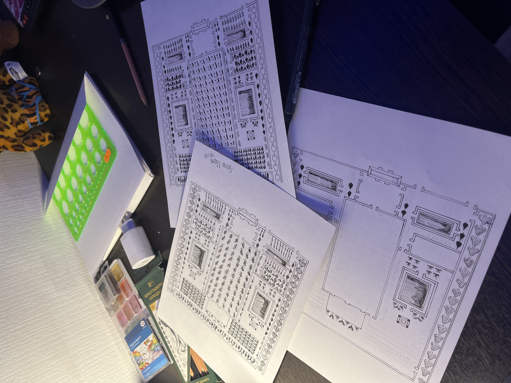 The image shows a workspace with several items related to architectural or garden design. There are three sheets of paper with hand-drawn designs that seem to be plans for a formal garden or landscaped area. The designs include geometric layouts, possibly representing planting beds, paths, and structures like pavilions or fountains. One of the drawings is partially gridded.
Other items include a pencil, a black pen, a stencil with circular cutouts, a set of watercolors, glue, and a box of graphite pencils. A plush toy with an animal print is also visible. The paper with the drawings on it is marked "Гусева Мария" and "гр. 200," indicating the creator's name and a group number. The drawings also have what looks like “09.2023” written on them. These marks could refer to the student's name, class, and a project or drawing date, respectively. No other identifying information is readily available from the image, so it's not possible to specify a location in Saint Petersburg, Russia. The context suggests an academic setting related to design or architecture.