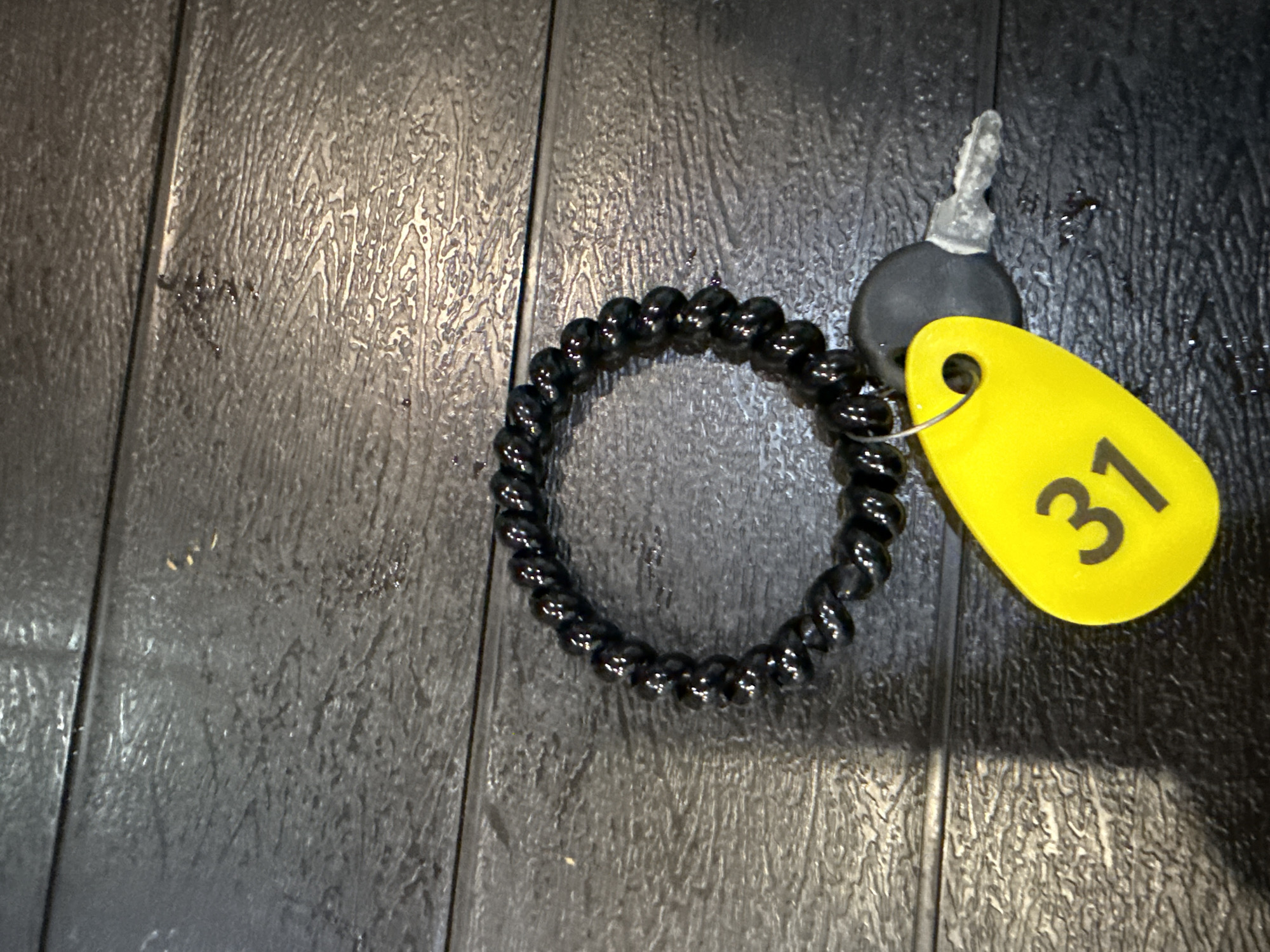 The image captures a close-up view of several objects resting on a dark, textured surface. The primary items include a black spiraled hair tie or bracelet made of numerous small, glossy beads. Adjacent to it is a single key with a black plastic head, to which a bright yellow oval key tag is attached by a metal ring. The number "31" is prominently printed in black on the yellow tag.
The underlying surface is dark, appearing to be made of planks or panels, evidenced by visible seams. It features a distinct ripple or wood-grain like texture and possesses a reflective sheen, suggesting it might be wet or have a glossy finish. No people or activities are depicted, and the objects are stationary. While the location context is Tobolsk, Russia, there are no specific visual cues within the image itself to indicate this geographical location.