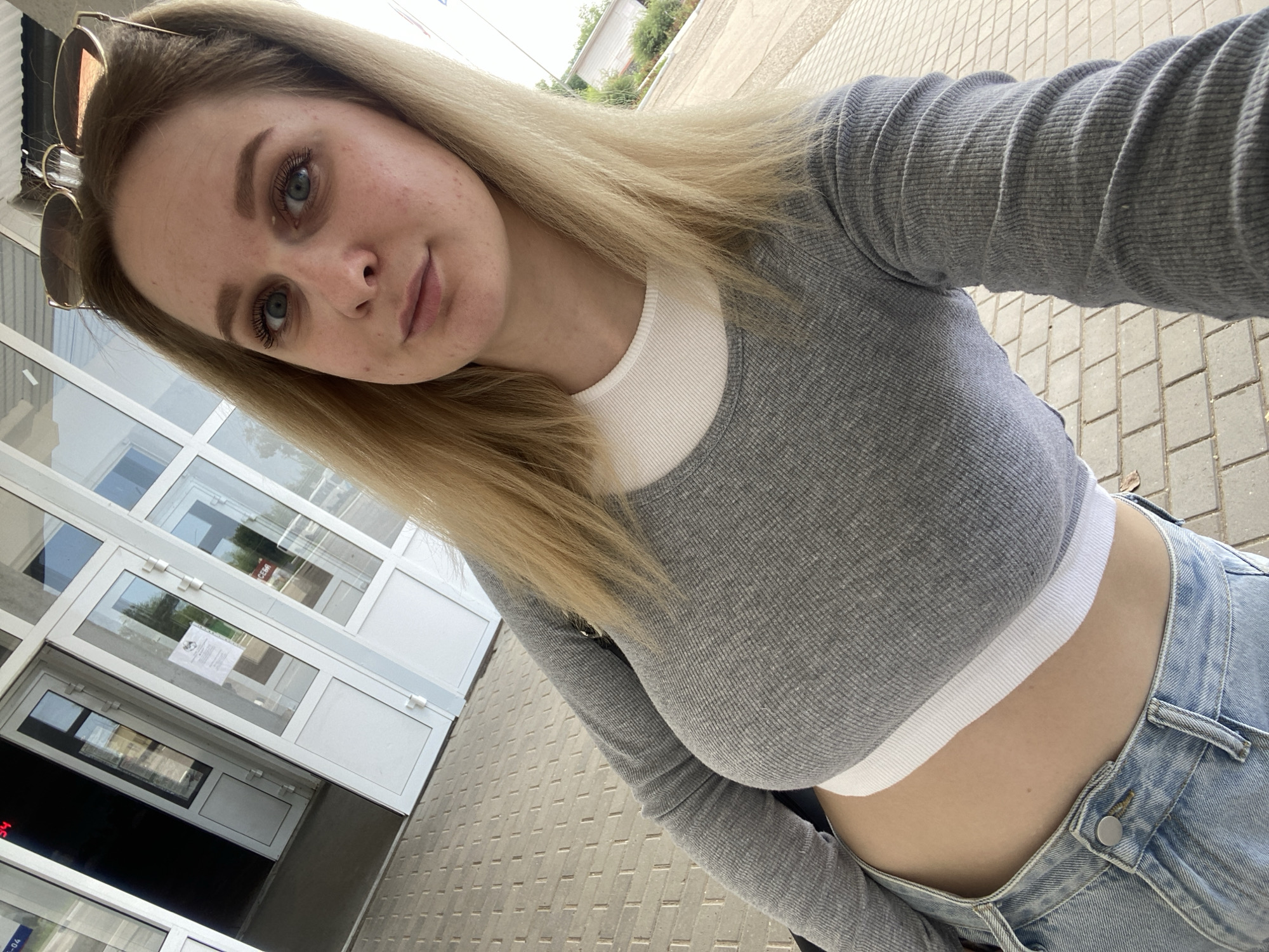 A young woman with blonde hair and blue eyes is taking a selfie outdoors. She is wearing a grey long-sleeved crop top over a white tank top and light blue jeans. She has sunglasses perched on top of her head. The background shows a building with glass doors and a paved walkway. The image appears to have been taken in front of a building, possibly an educational institution or a public facility, located in Chaikovsky, Russia. The lighting suggests it is daytime.